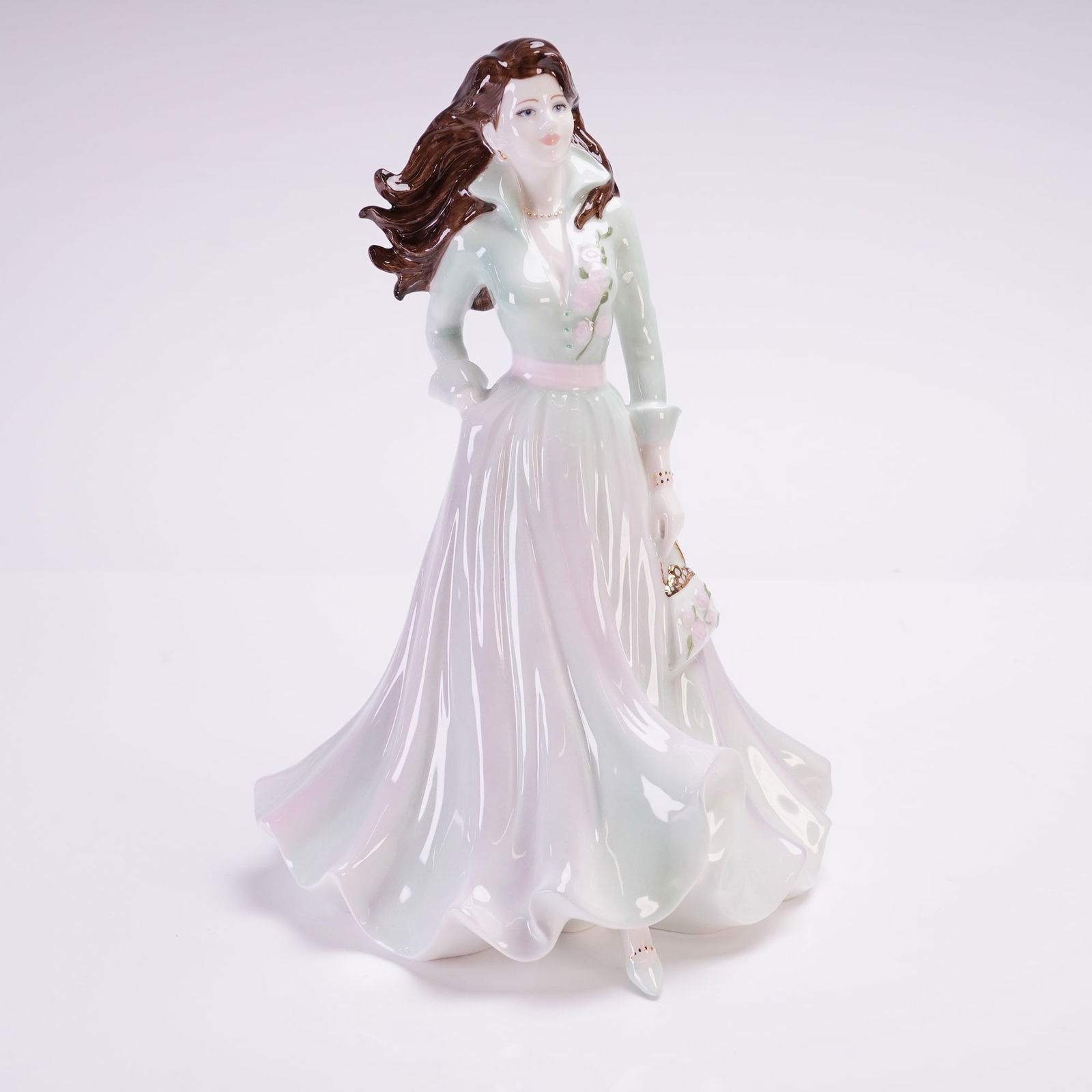 Coalport Ladies of Fashion Figurine, Sarah: This hand-decorated piece depicts a woman in a flowing, full-length gown of pale green and lavender hues with floral accents. The figure is styled with long, wind-swept brown hair and carries a small