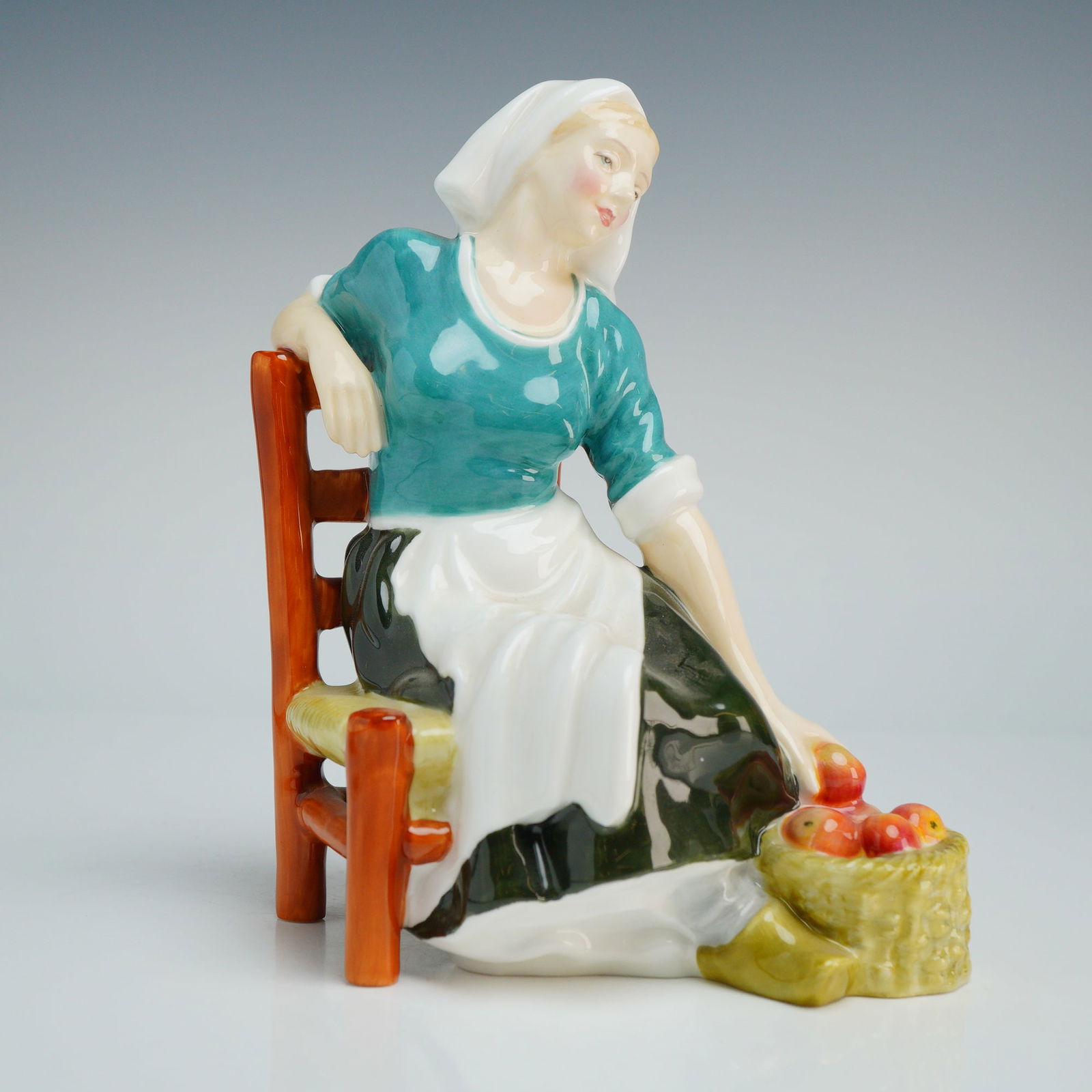 Royal Doulton Figurine, Apple Maid HN2160: The piece depicts a seated woman in traditional attire, featuring a teal blouse, white apron, dark green skirt, and a white headscarf. The figure is posed leaning against a rustic wooden chair, reachi