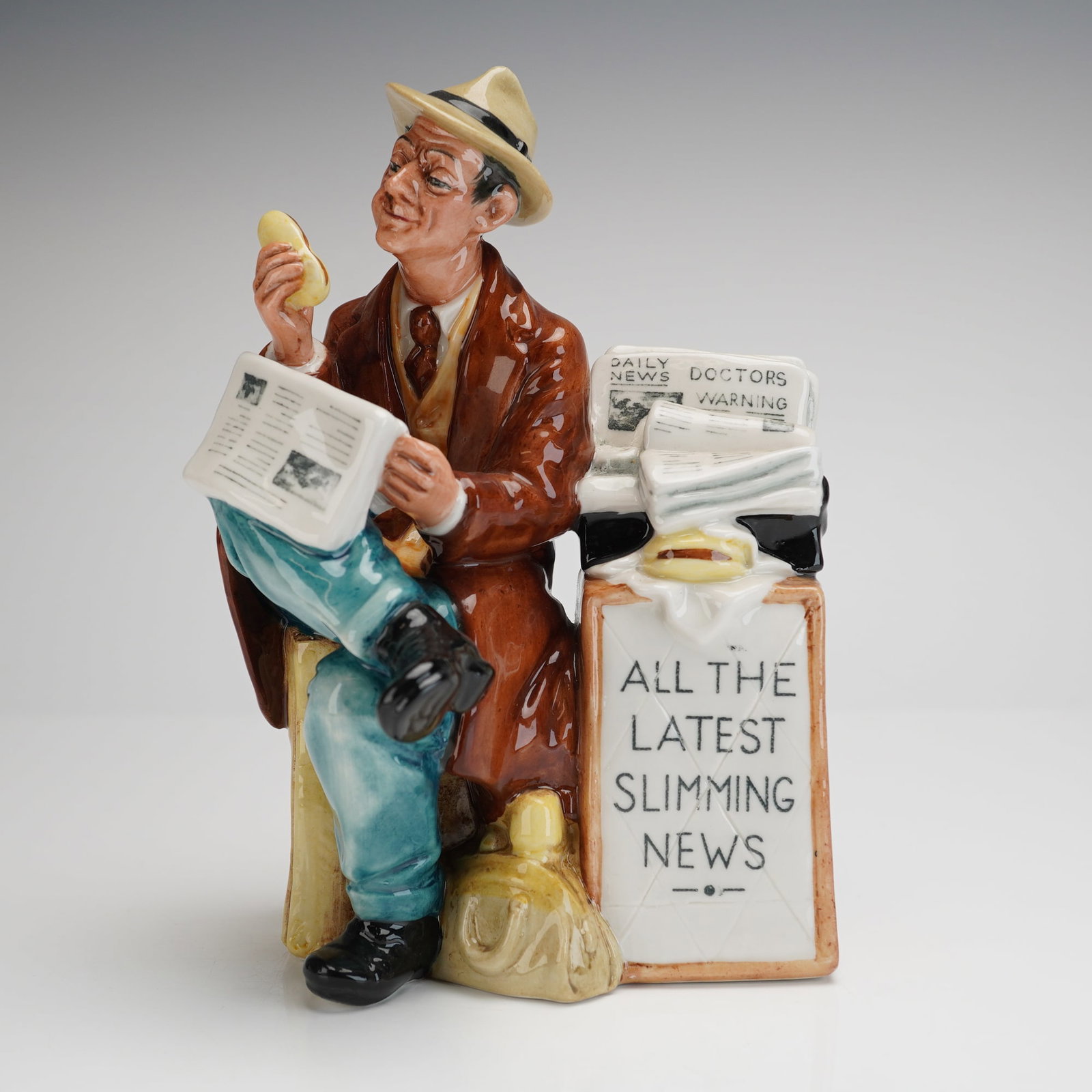 Royal Doulton Figurine Stop Press HN2683: This hand-painted porcelain figure depicts a seated man, typically associated with a newspaper vendor or reader, dressed in a brown trench coat and fedora. The man is shown holding a yellow, banana-sh