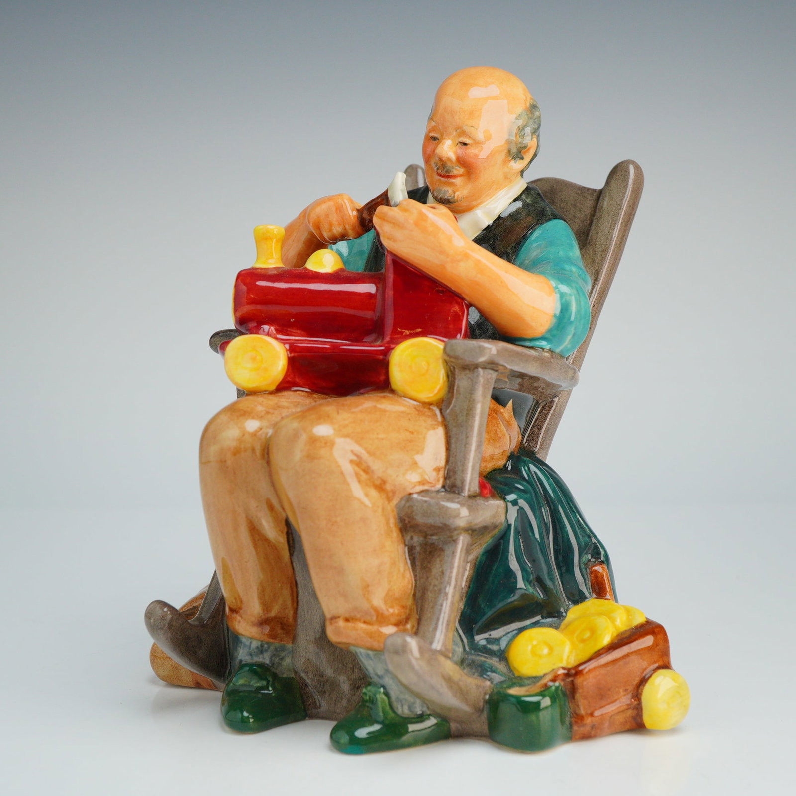 Royal Doulton Figurine, The Toymaker HN2250: This Royal Doulton depicts an elderly craftsman seated in a gray-toned rocking chair. The figure is shown assembling a red toy steam locomotive and is dressed in a turquoise shirt and a dark vest over