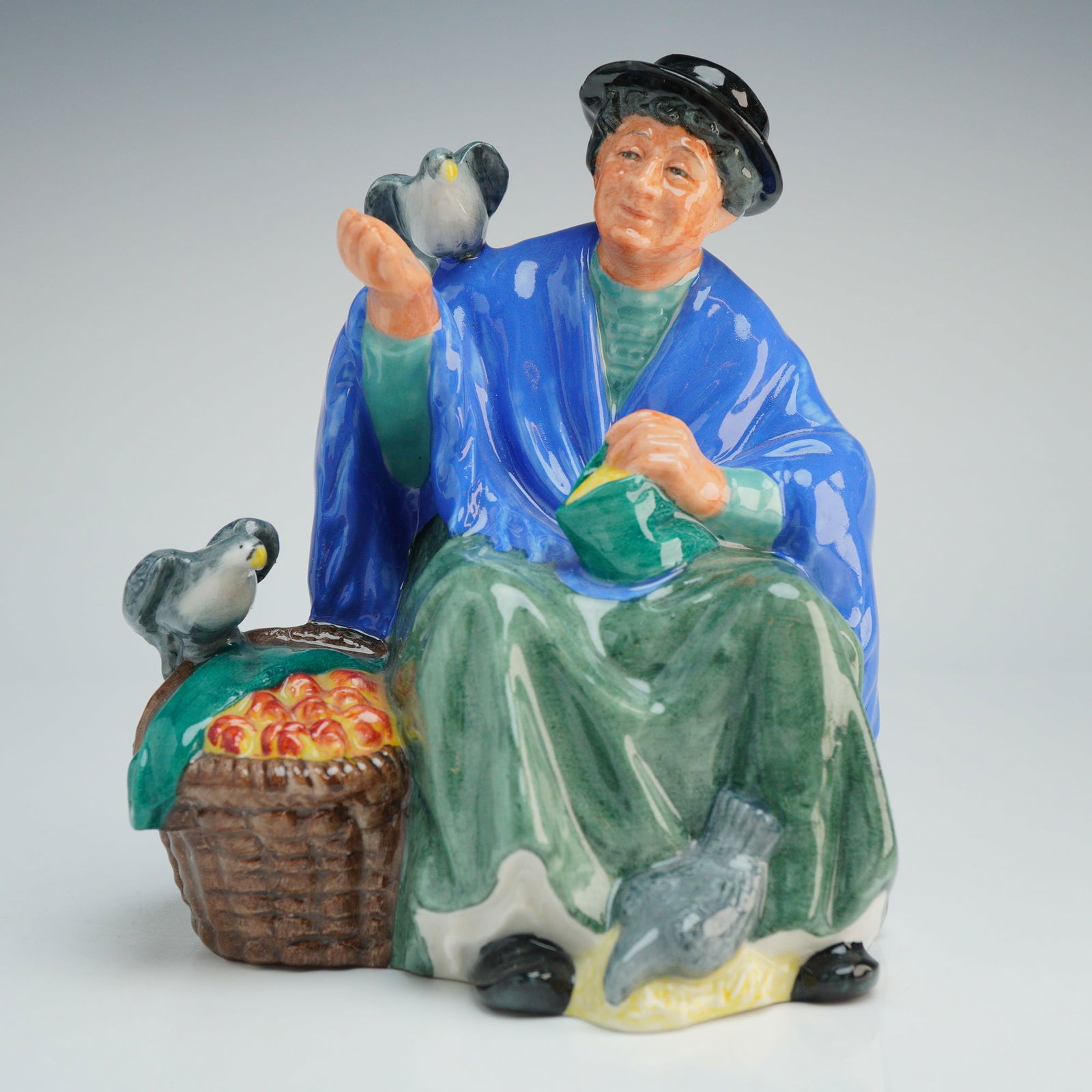 Royal Doulton Figurine, Tuppence A Bag HN2320: The figure depicts an elderly woman seated upon a gray stone wall feeding pigeons. She is wearing a blue shawl over a green skirt and blouse with a black hat. A woven basket containing orange fruit is