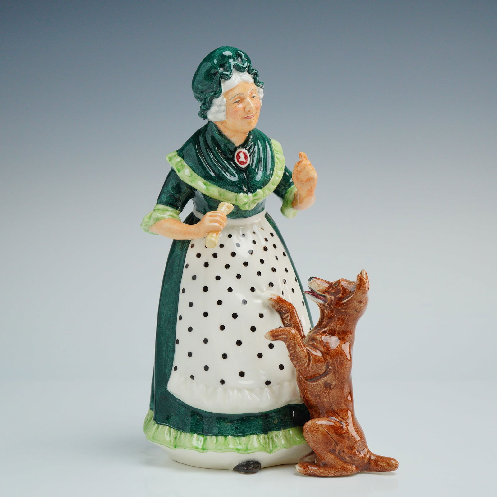 Royal Doulton Porcelain Figurine, Old Mother Hubbard HN2314: This Royal Doulton porcelain figurine, titled Old Mother Hubbard (HN 2314), is a hand-painted glazed ceramic work. The piece depicts an elderly woman wearing a tiered dark green dress with light green