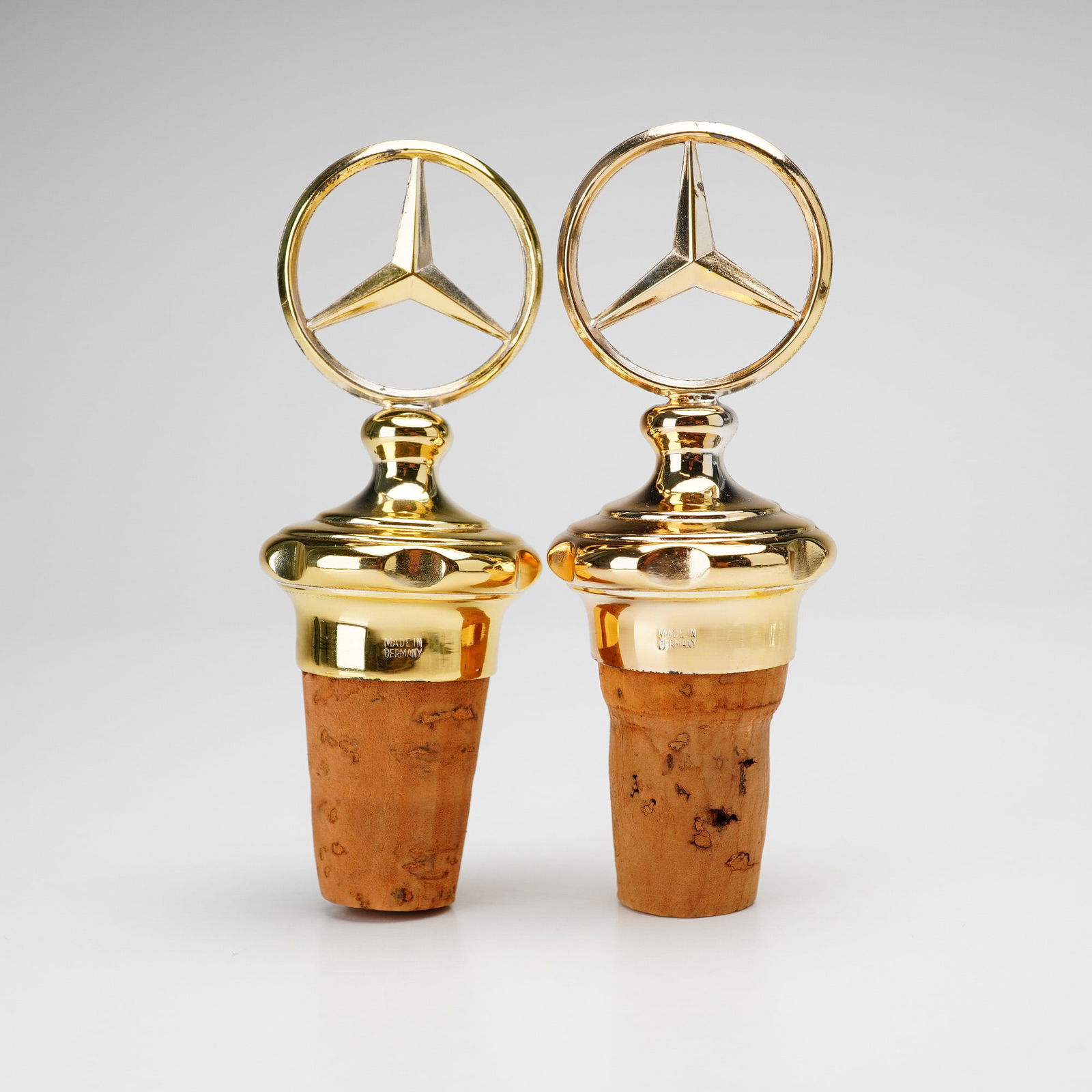 Pair of Mercedes Benz Gold-Tone Wine Bottle Stoppers: Two gold-toned Mercedes Benz emblem tops cork base. Made in Germany. Original boxes are included. Dimensions: 1.25"L x 1.25"W x 3.50"H Weight: 2.60oz