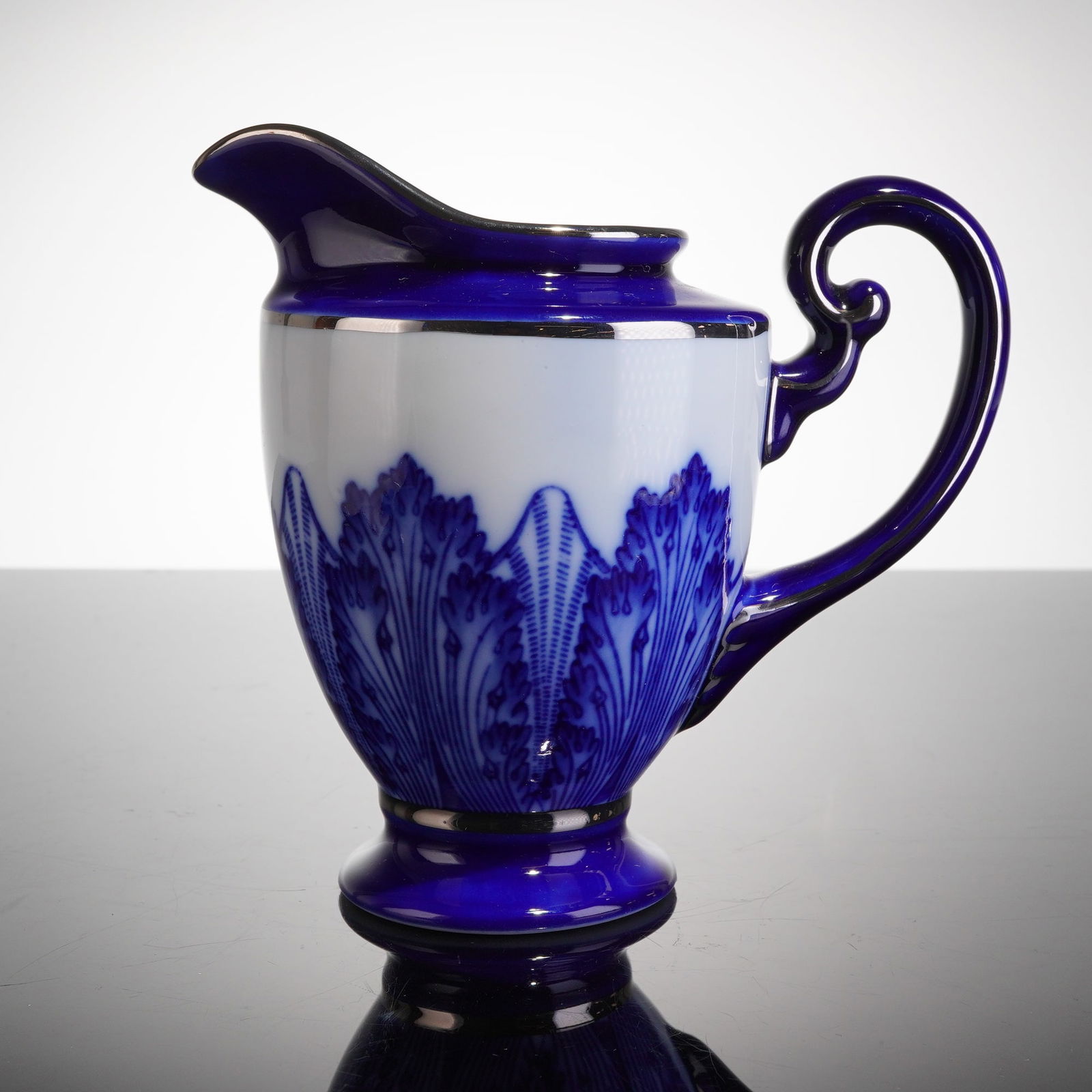 Bombay Company Blue and White Porcelain Creamer: The creamer has an hourglass-like body with a flaring foot, executed in white and deep cobalt blue glaze. The lower half is decorated with intricate blue transfer-printed stylized repeating leaf or fr