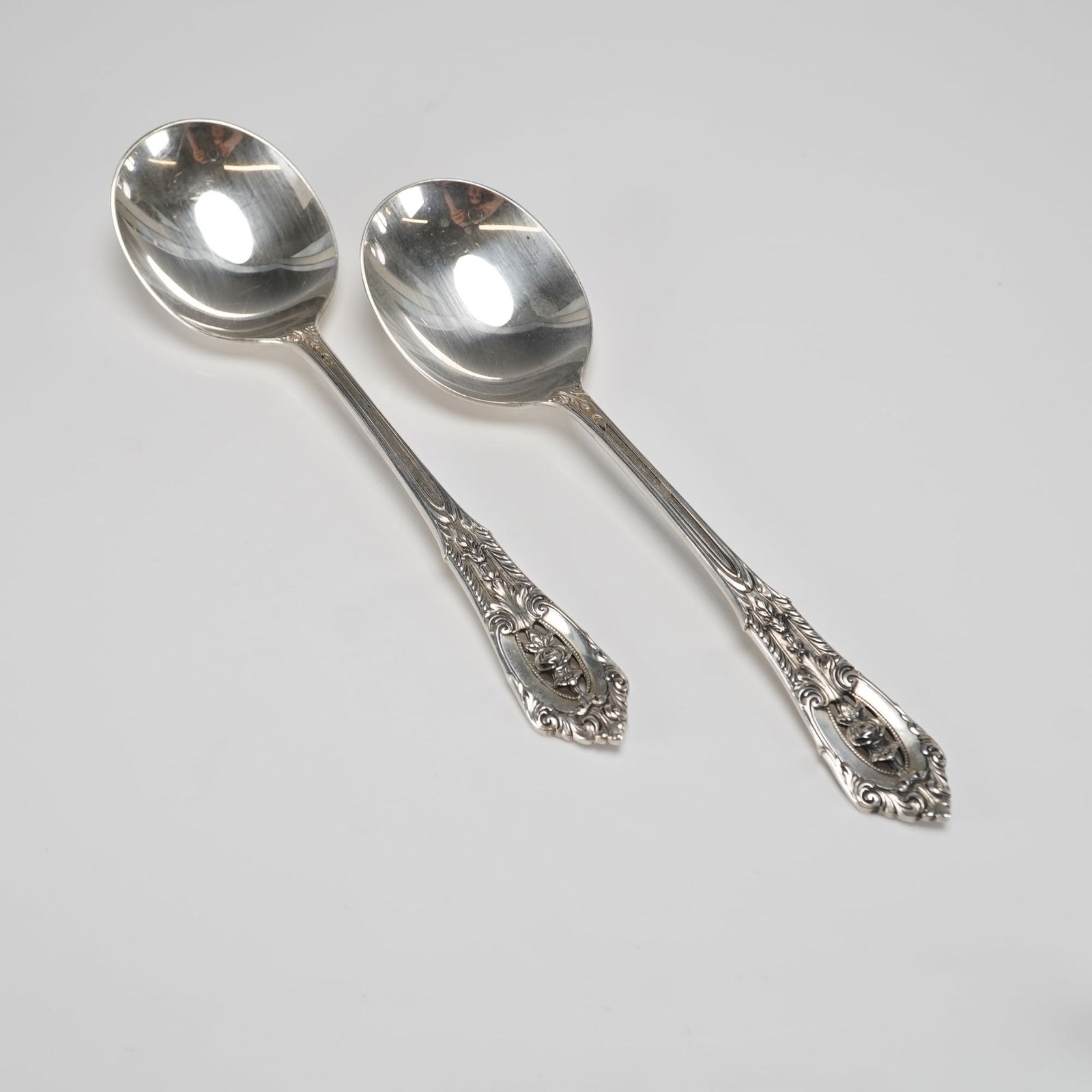 A Pair of Wallace Rose Point Sterling Silver Cream Soup Spoons: A pair of Wallace Rose Point sterling silver cream soup spoons. These two sterling silver soup spoons feature a highly ornate Rococo Revival style, characterized by elaborate scrolling relief, acanthu