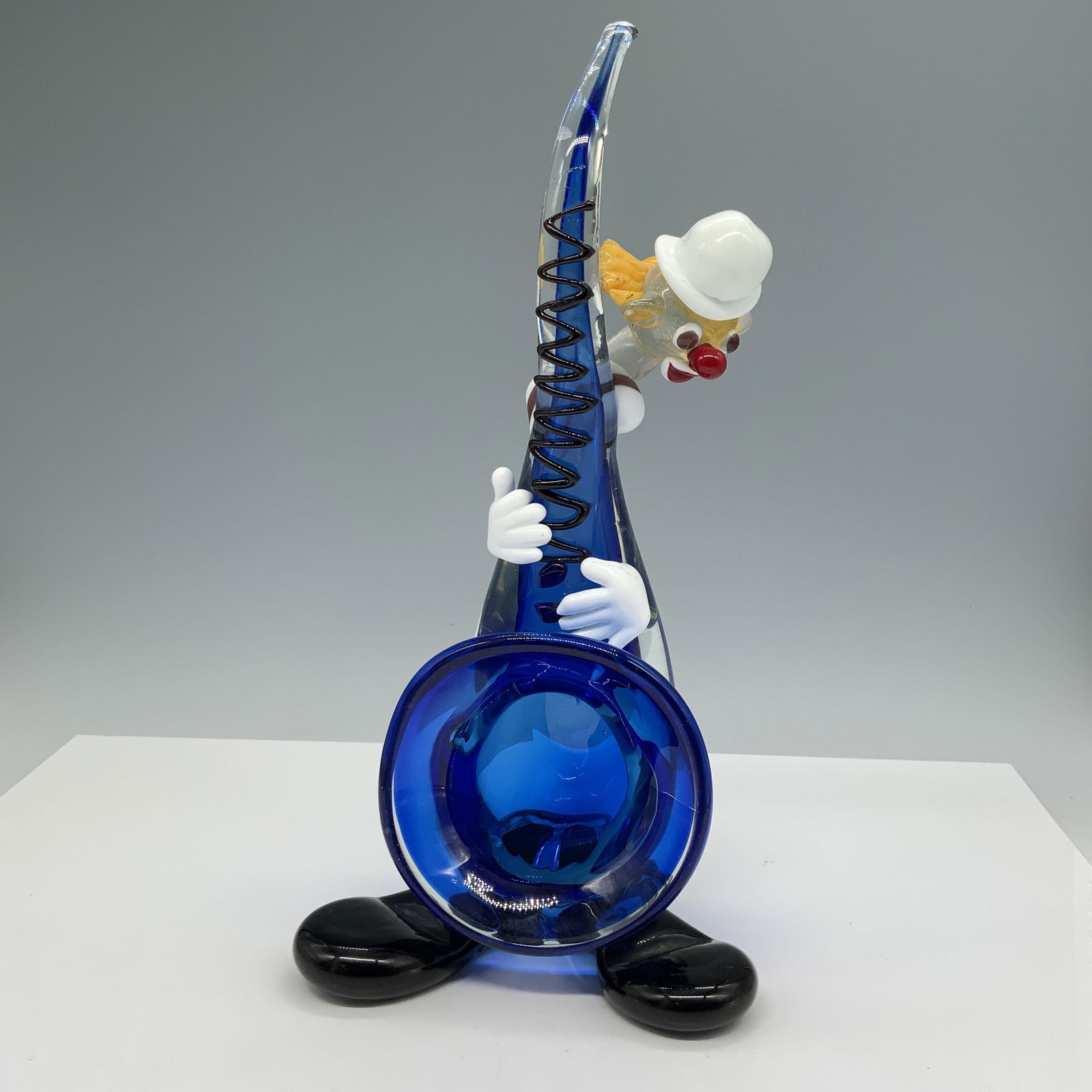 Blue Horn Glass Clown Figurine: This playful glass figurine features a clown holding a blue horn, with vibrant detailing. The clown is carefully crafted with yellow and white hair, a red nose, and black shoes, providing a cheerful e
