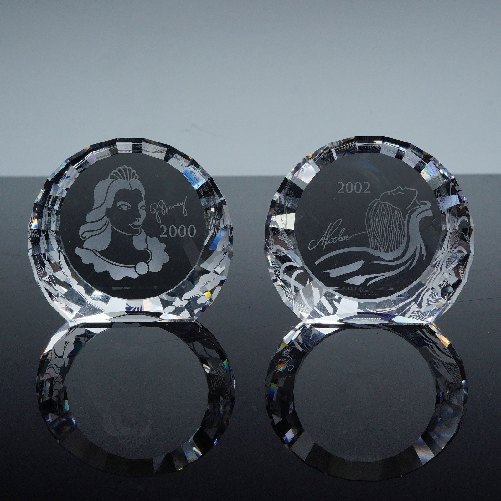 2pc Swarovski Crystal 2000 Columbine & 2002 Isadora Event Paperweight: Pair of Swarovski crystal event paperweights including the 2000 Columbine design featuring an etched profile of the Columbine masquerade figure, and the 2002 Isadora design with stylized dancer engrav