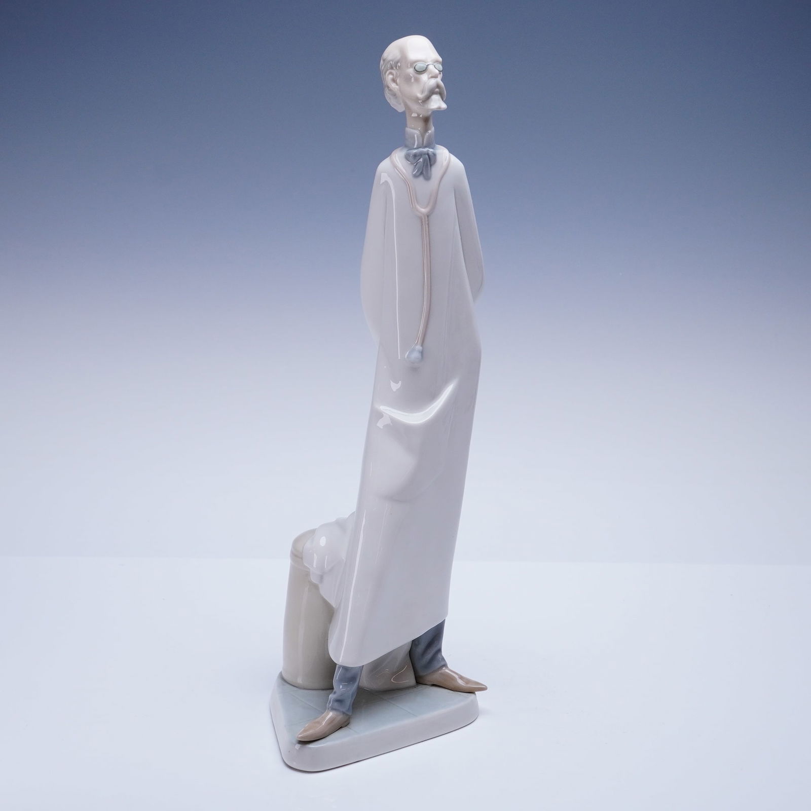 Lladro Porcelain Figurine The Doctor Medico 1004602: This Lladro porcelain figurine depicts an elderly male physician characterized by a prominent mustache, wearing a white lab coat, and carrying a stethoscope around his neck. The figure is posed standi