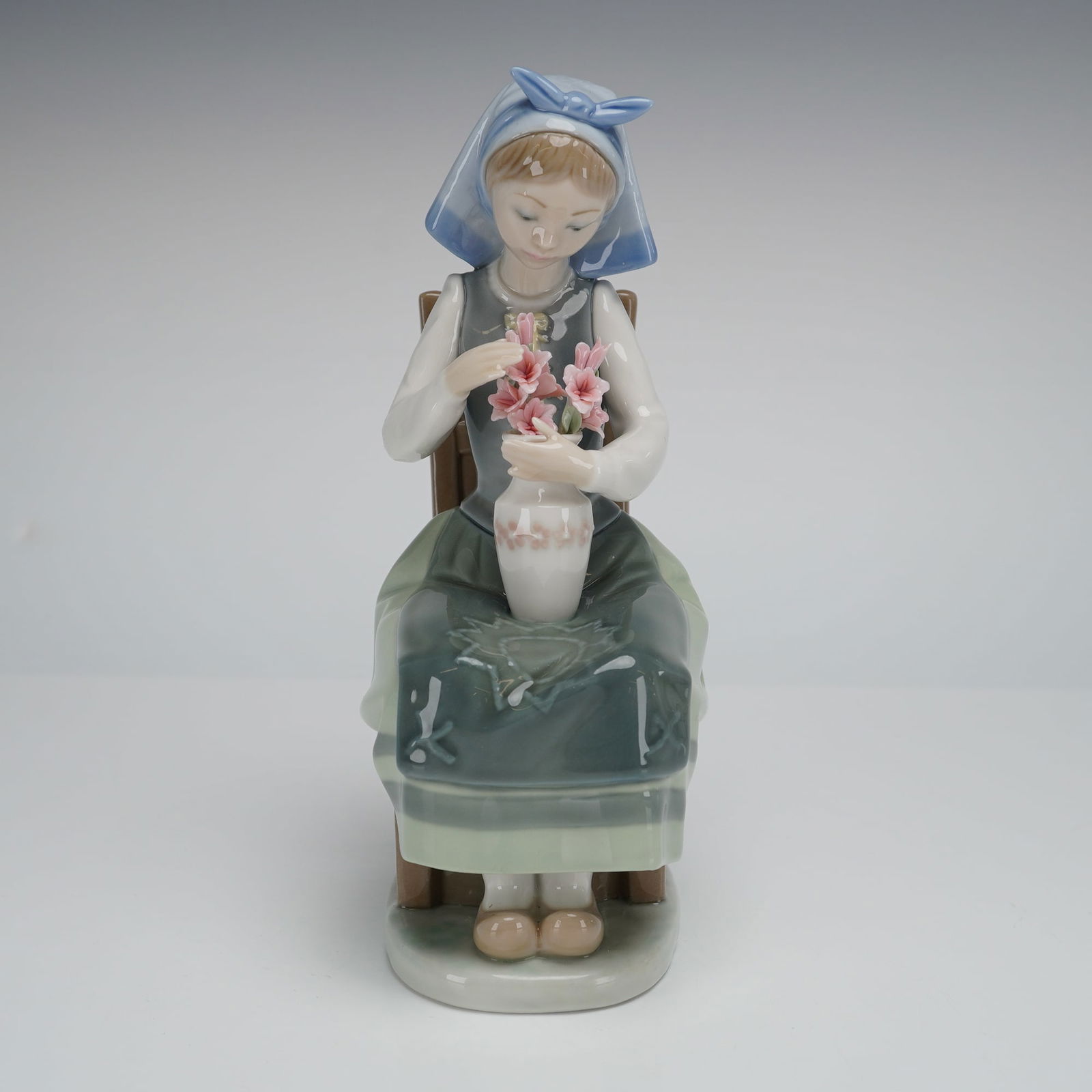 Lladro Flower Harmony Porcelain Figurine 1001418: A glazed porcelain figurine by Lladro, titled "Flower Harmony," model number 1418. The piece depicts a young female figure seated on a wooden chair, engaged in arranging pink flowers into a white vase