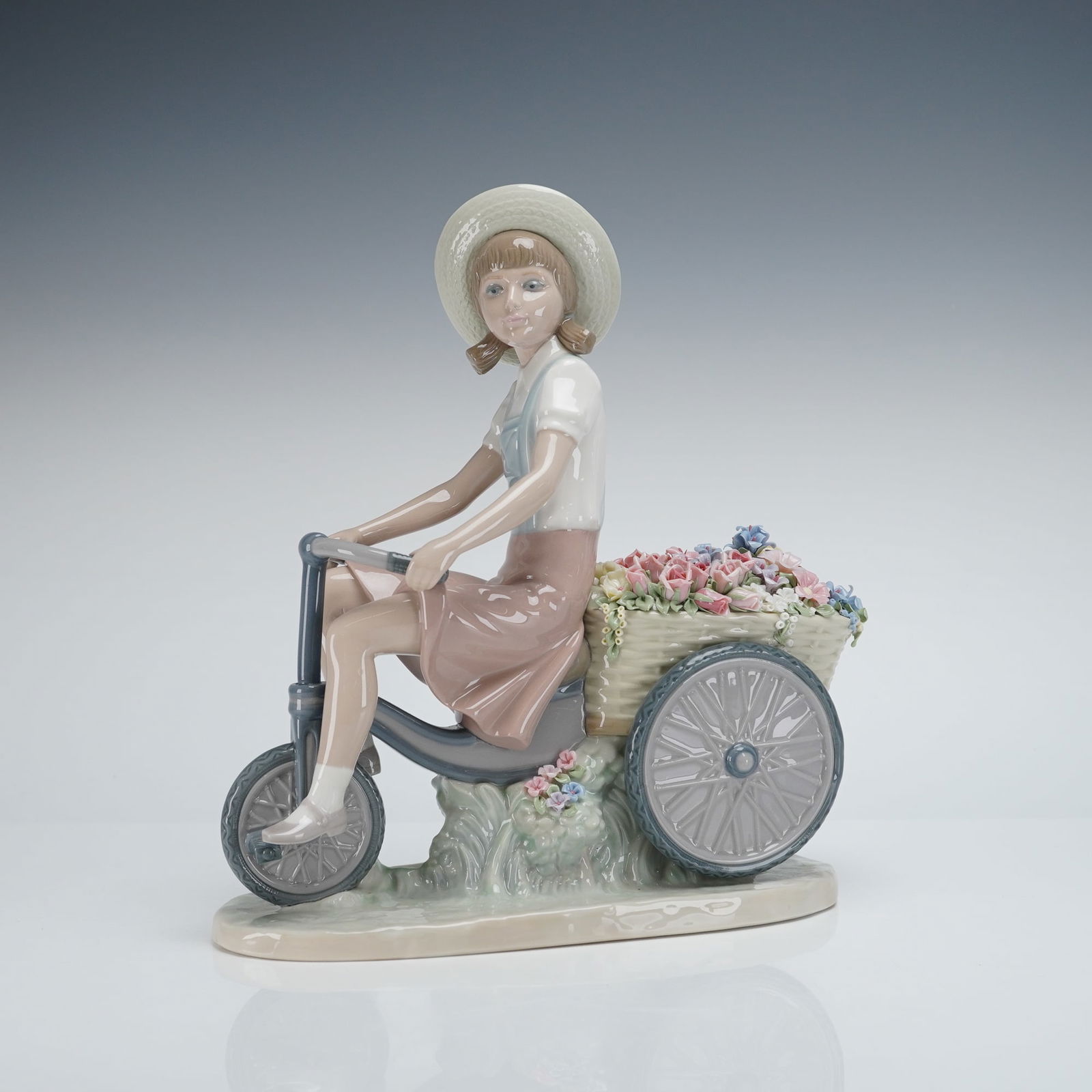 Lladro Girl with Flowers in Tow Figurine 1005031: The glazed Lladro porcelain figurine depicts a young female figure wearing a sun hat and pastel clothing, riding a three-wheeled delivery bicycle. The rear of the bicycle features a woven basket dense