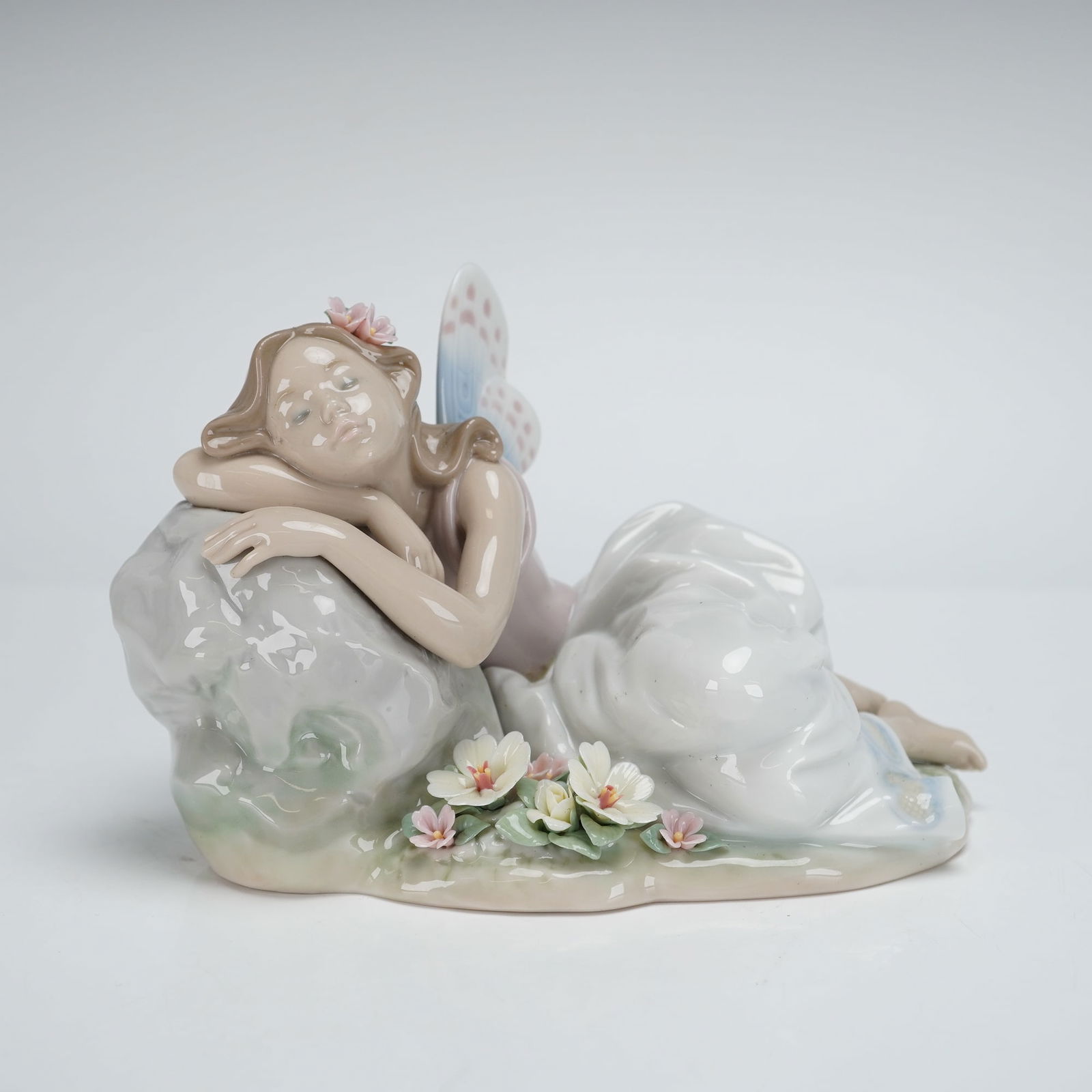 Lladro Princess of the Fairies Figurine 7694: A Lladro Porcelain Figurine, titled Princess of the Fairies, model number 7694. The glazed porcelain piece depicts a reclining nymph or fairy figure resting her head upon a gray rock formation. The fi