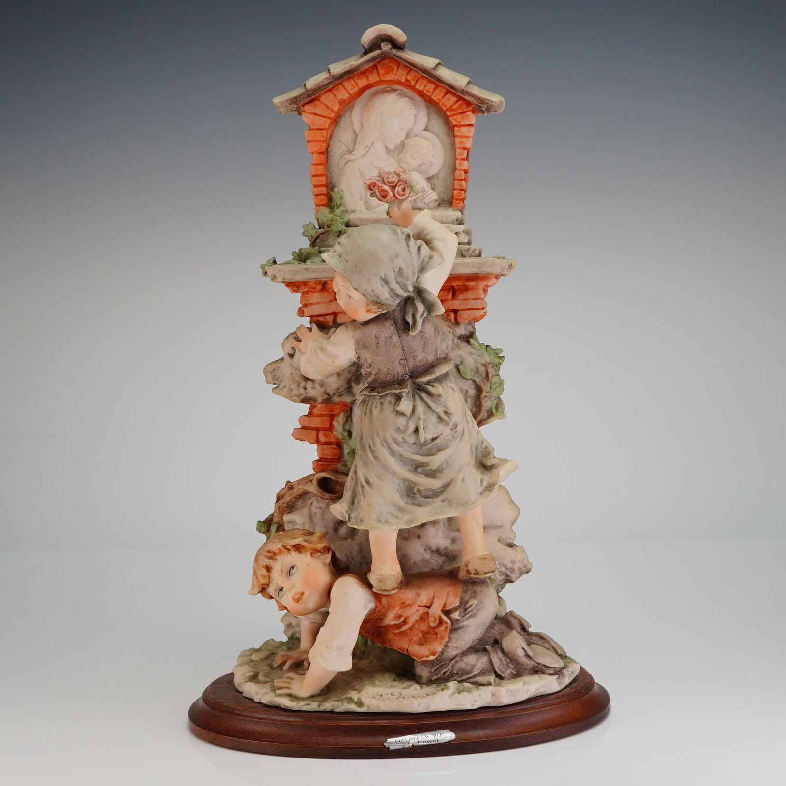 Giuseppe Armani Gulliver's World Flowers for the Shrine Sculpture: The highly detailed figurative sculpture depicts two children interacting with a roadside shrine: a girl, dressed in period clothing and a bonnet, is climbing onto the back of a crouching boy to place