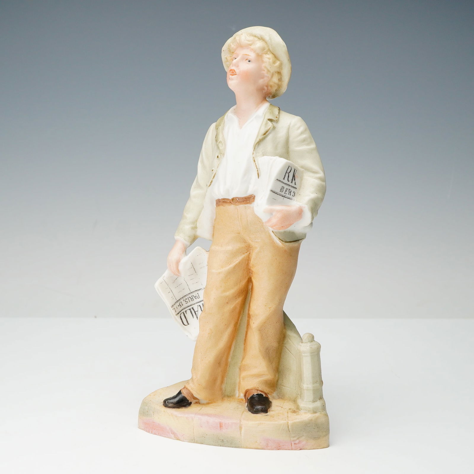 Gebruder Heubach Bisque Porcelain Newspaper Boy Figurine: This is a single bisque porcelain figurine attributed to Gebruder Heubach, depicting a newsboy or street vendor. The figure is rendered in a naturalistic style, wearing a cap, light jacket, and trouse