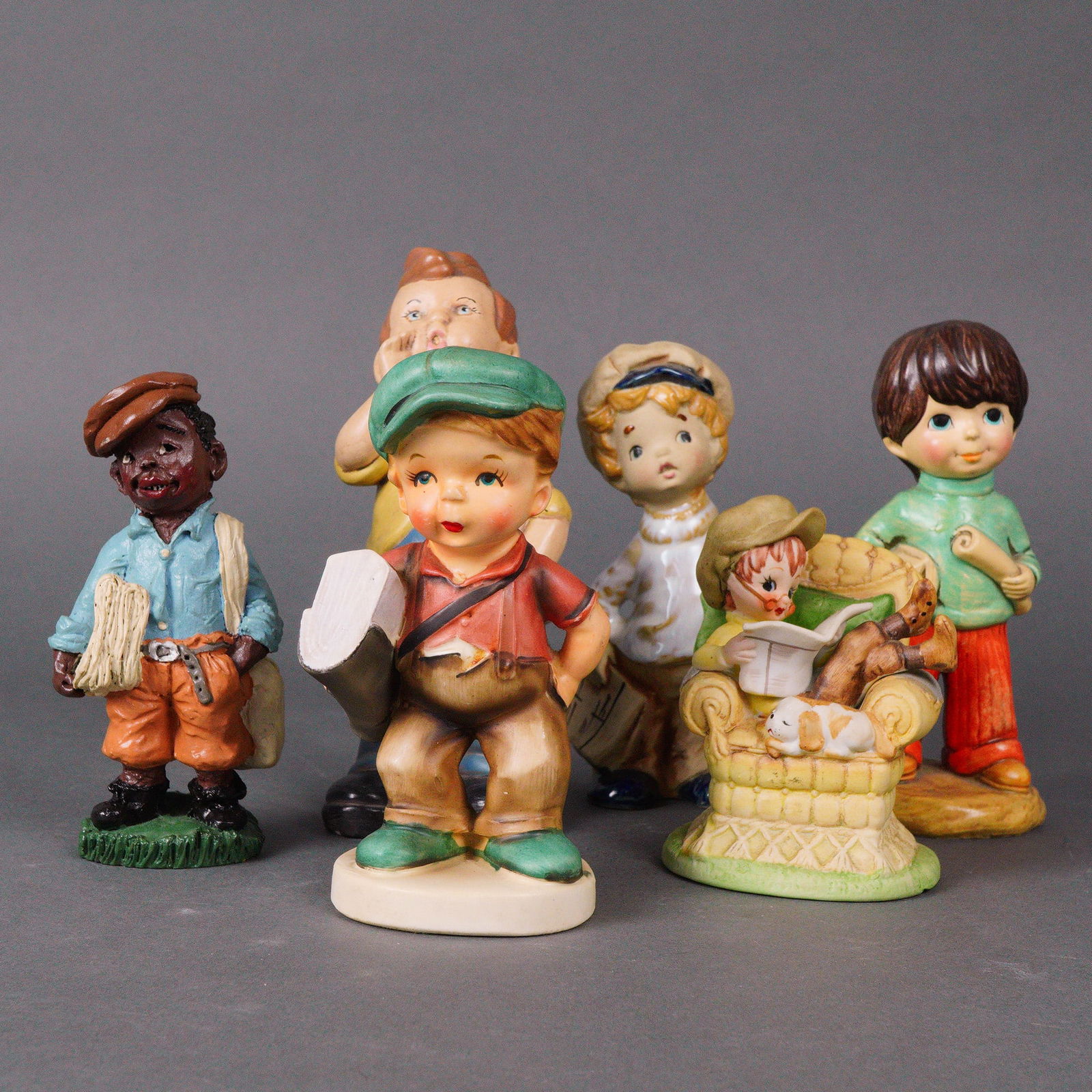A Collection of Six Glazed Ceramic Newsboys Figurines: A group of six vintage figurines depicting children with newspapers in various poses. The figures are made from painted ceramic or composite materials and show a range of styles from cartoonlike to mo
