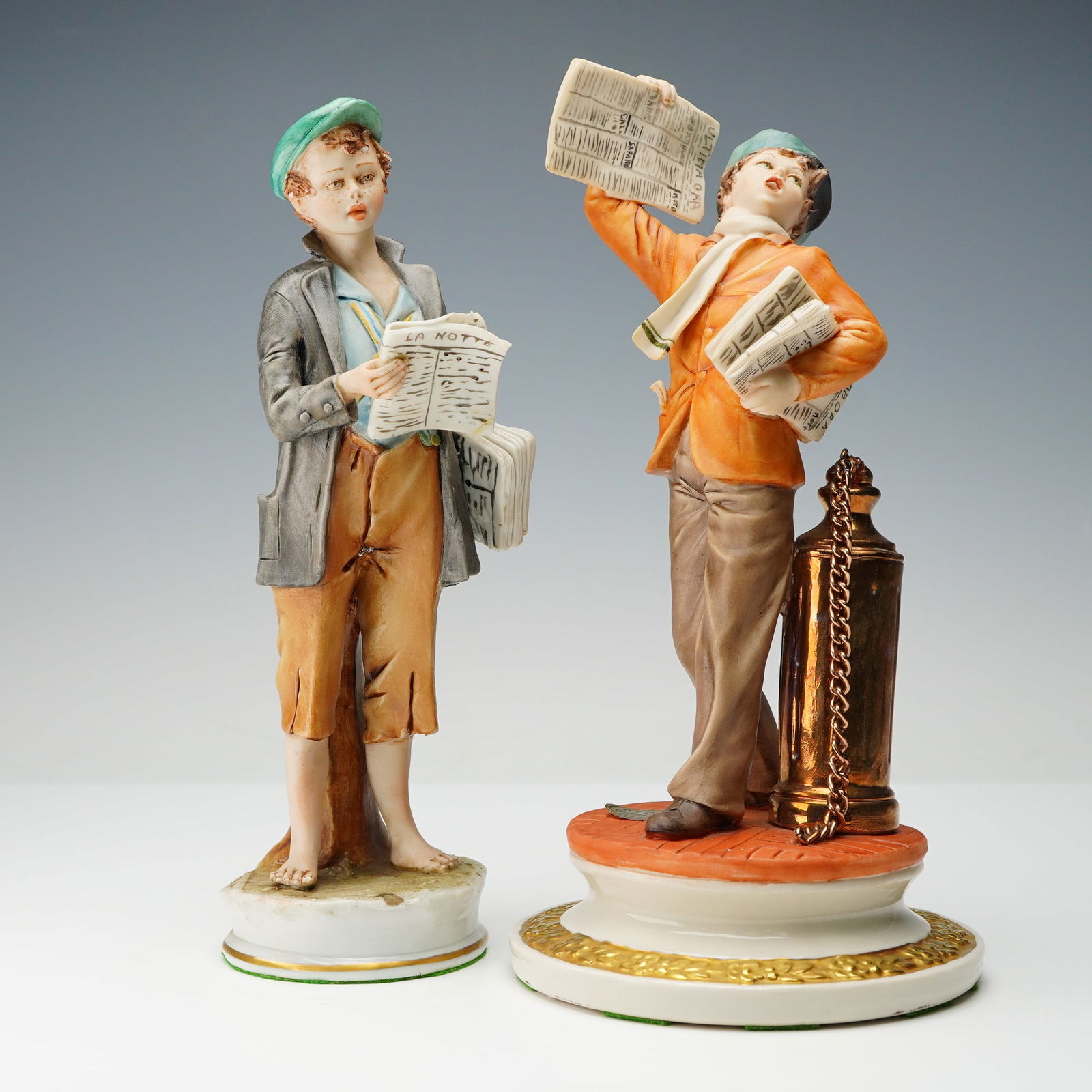2pc Capodimonte and Sandro Maggioni Porcelain Newspaper Vendor Figurines: A pair of polychrome glazed porcelain figurines, depicting two young male newspaper vendors. The figures are hand-painted with detailed clothing and facial expressions. The figure on the left holds pa