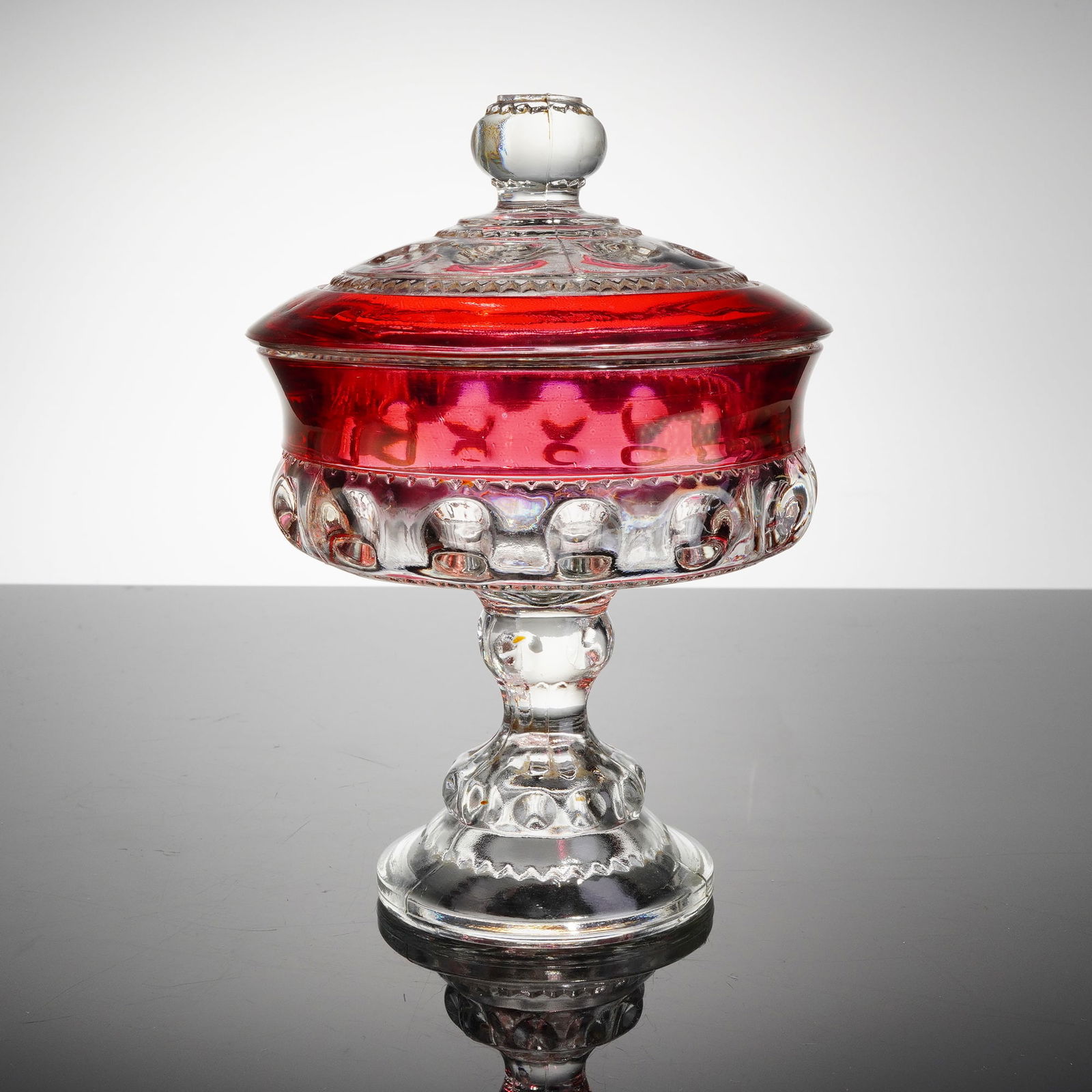 Crown Ruby Glass Footed Candy Dish: The covered dish is fabricated from heavy clear pressed glass, featuring a deep ruby red overlay or flashing applied to the upper bowl and fitted lid sections. This piece functions as a pedestal candy