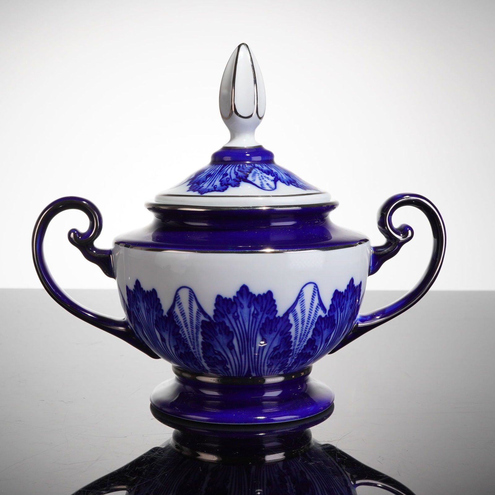 Bombay Company Blue and White Lidded Sugar Bowl with Silver Trim: The sugar bowl is executed in a Neoclassical style, featuring a cobalt blue and white color scheme. The two-handled urn form rests on a pedestal foot. The lower white reserve band is decorated with