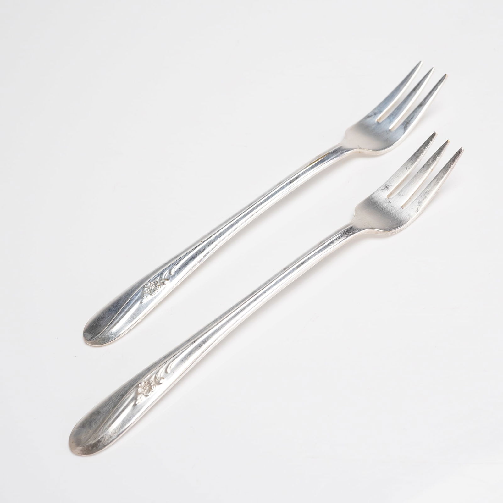 A Pair of Holmes & Tuttle Meadow Flower Silverplate Cocktail Forks: A pair of silver-plated cocktail or dessert forks manufactured by Holmes & Tuttle Manufacturing Company (H. & T. MFG. CO.) in the Meadow Flower pattern. Each piece is constructed of