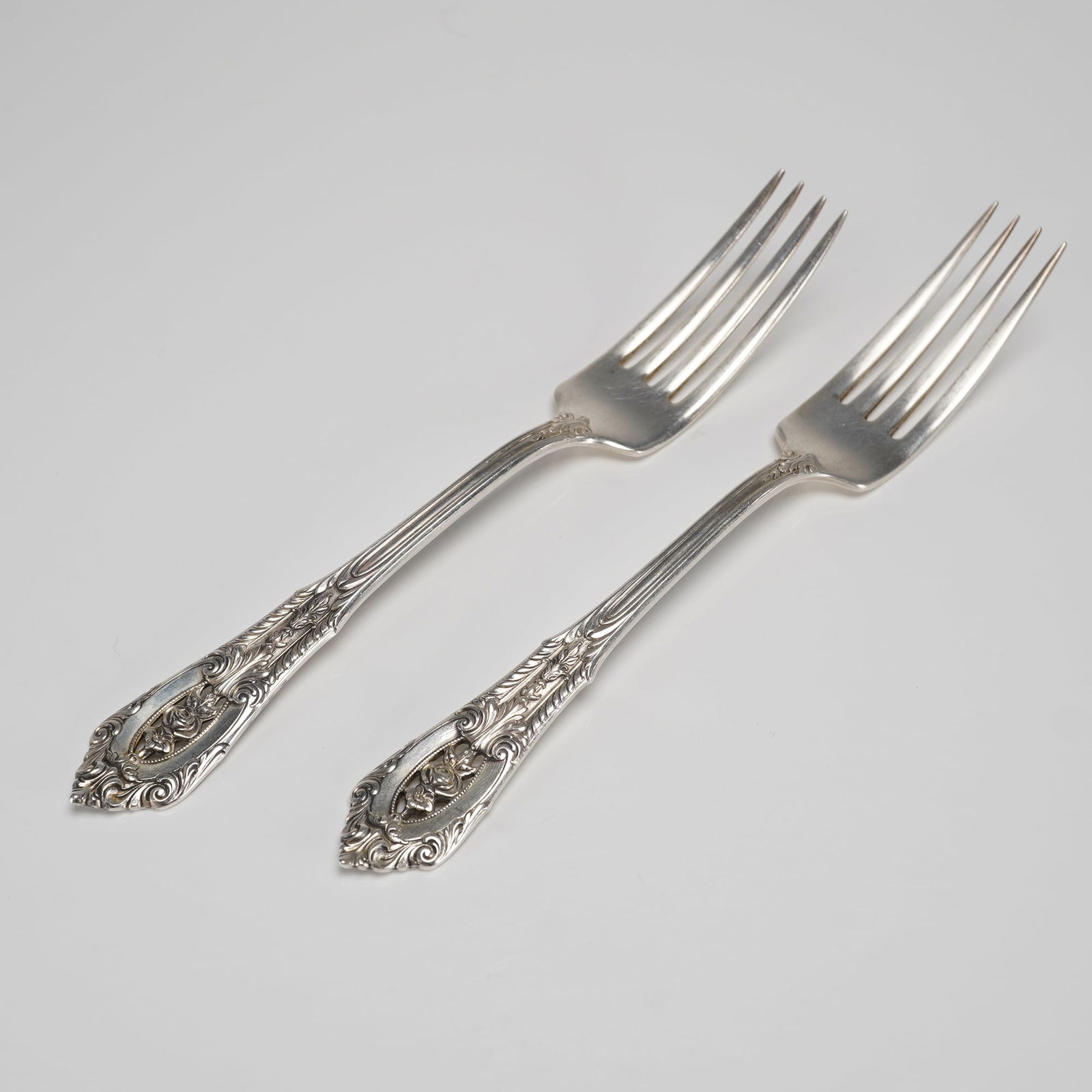 A Pair of Wallace Rose Point Sterling Silver Dinner Forks: A pair of sterling silver forks manufactured by Wallace in the Rose Point pattern. These two sterling silver forks feature a highly detailed Rococo Revival style handle. The decoration includes dense