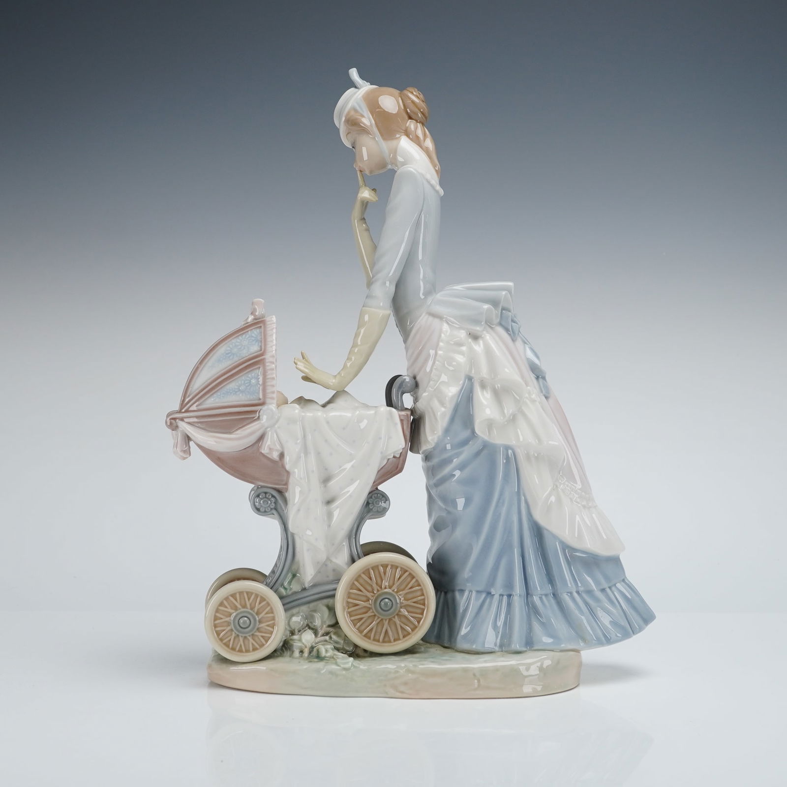 Lladro Babys Outing Figurine 4938: The figurine depicts a standing woman dressed in a period late 19th/early 20th-century style gown, executed in pale blue, pink, and white glazes, bending slightly to look down at a baby sleeping in a