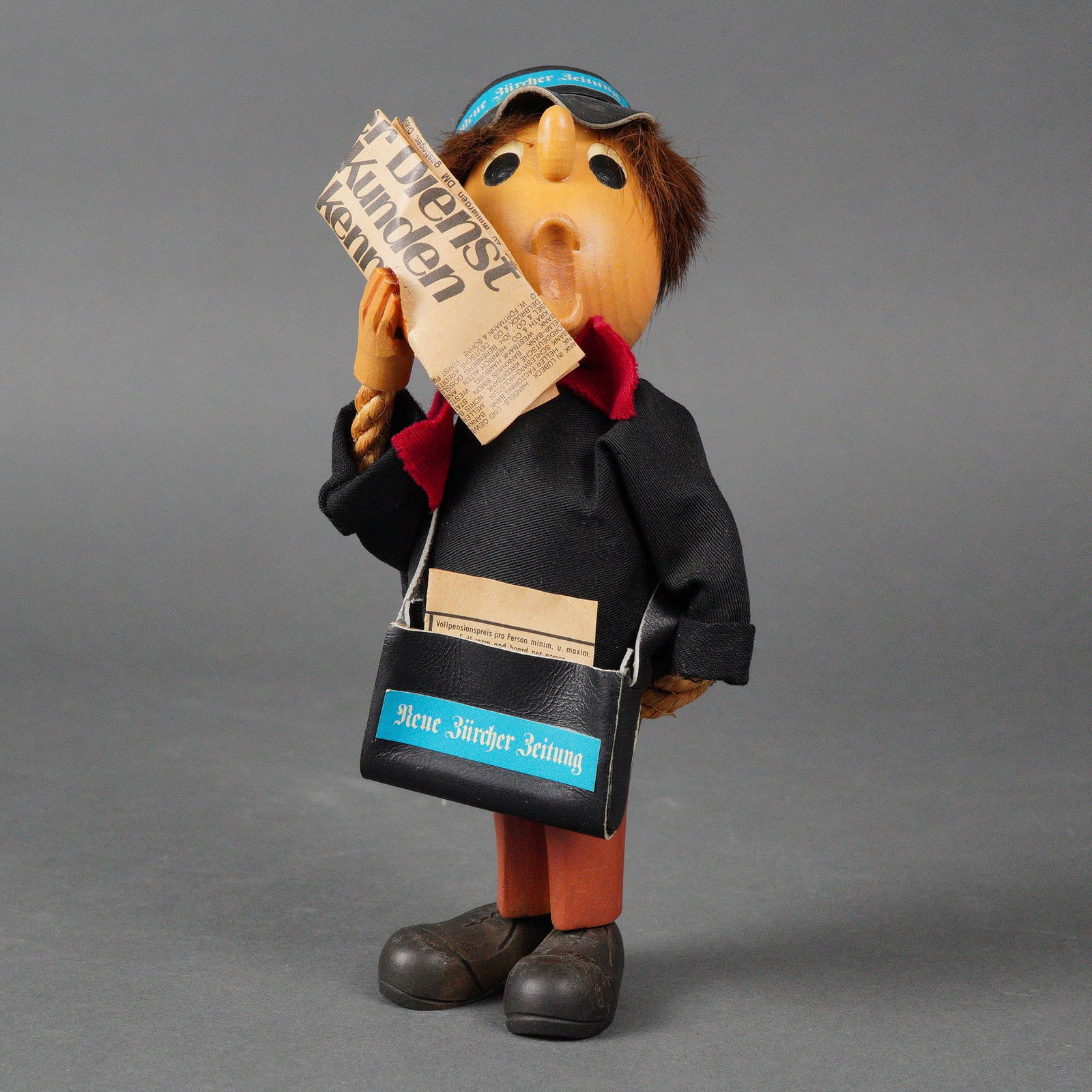 Anri Romer Casy Boys Carved Wood Newsboy Figurine: The figure features a carved wooden head with exaggerated features, hands, legs, and shoes. It is dressed in fabric clothing in black and orange, a red fabric scarf, and a black cap and shoulder bag,