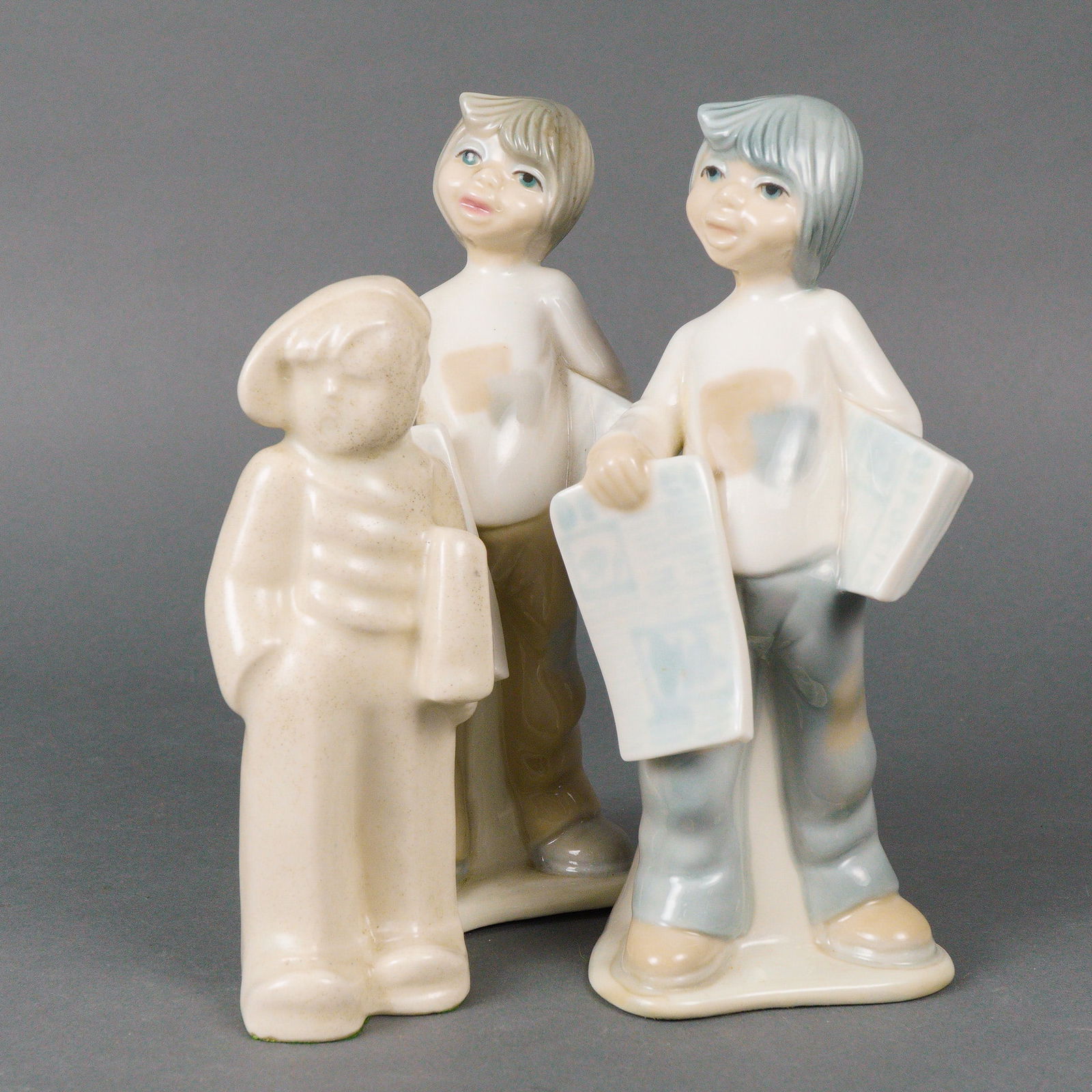 Three Tengra Ceramic Newspaper Boys Figurines: Three ceramic figurines depicting boys holding newspapers. Two glazed and hand painted figures are by Tengra of Spain, modeled in soft cream, blue, tan, and flesh tones with simplified features and ca