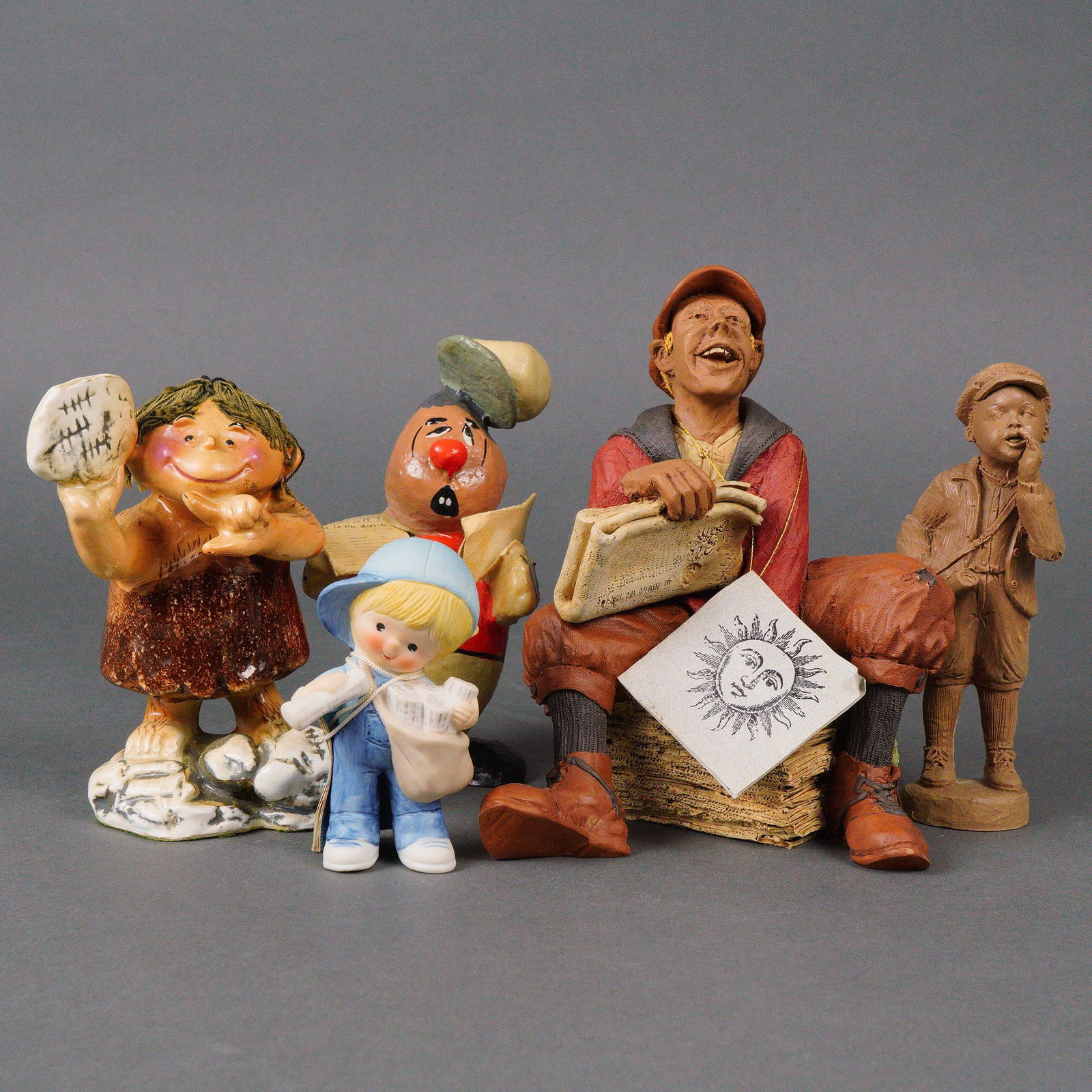 A Group of Five Newspaper Figurines: A group of five late 20th-century figurines, primarily crafted from painted resin or ceramic, displaying a whimsical, cartoonish, and expressive style. The group features a laughing news vendor seated