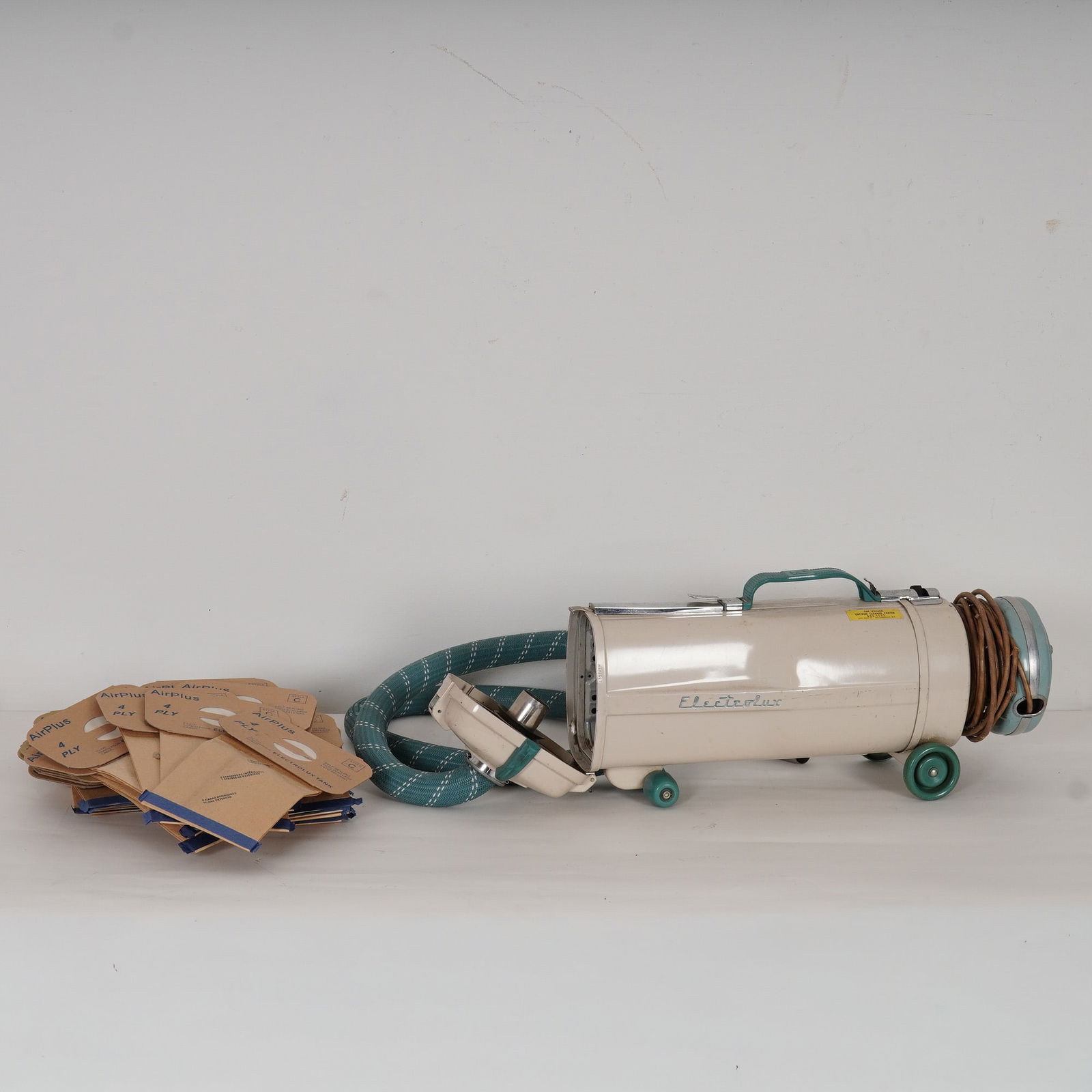 Electrolux Canister Vacuum Cleaner with Attachments and Bags: A mid-20th-century Electrolux canister vacuum cleaner, finished in cream with teal accents, accompanied by its flexible teal hose, metal wand, and assorted attachments. The cylindrical body sits on fo