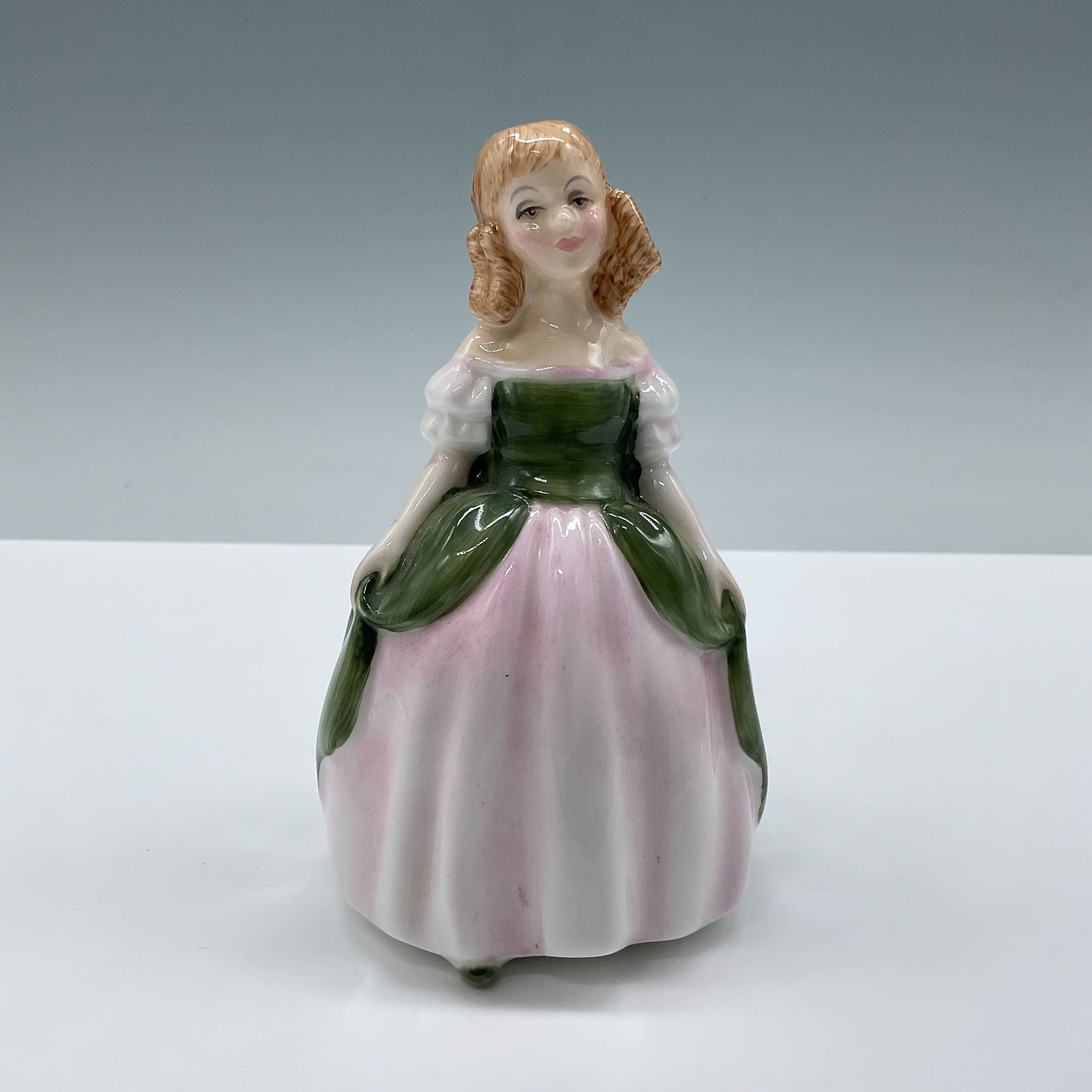 Penny HN2338 - Royal Doulton Figurine: A young girl in a lovely green and pink dress, her blonde curls framing her face, with her hands gently resting at her sides. Royal Doulton backstamp. Artist: Peggy Davies Issued: 1968 - 1995