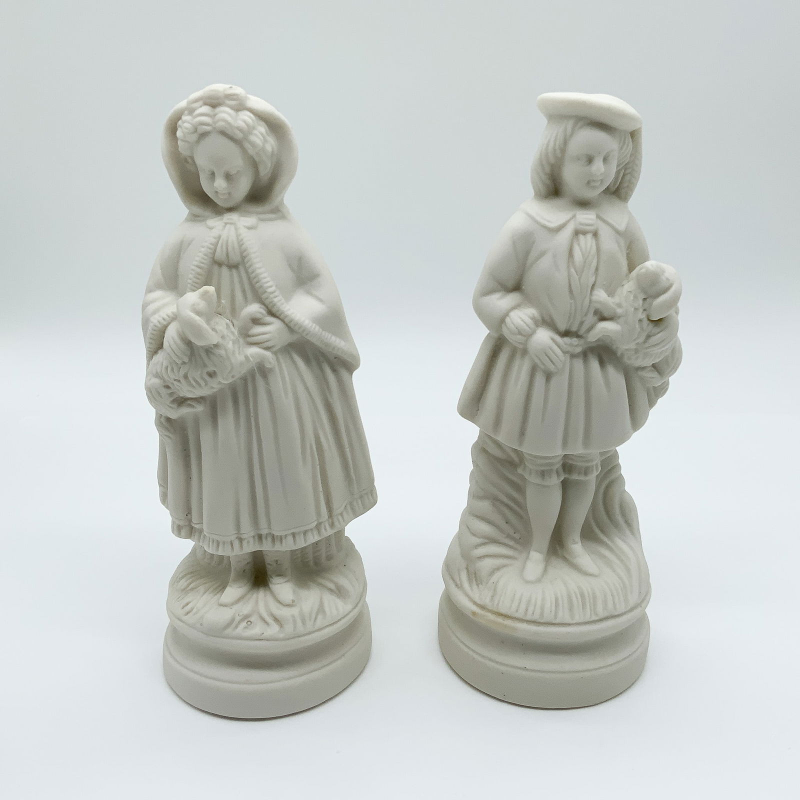 Pair of Bisque Porcelain Figurines: Pair of bisque porcelain figurines featuring a girl with a lamb and a boy with a bird. Detailed Victorian-style design with a matte finish. Unmarked bases, measuring 7.25H. Perfect for porcelain or
