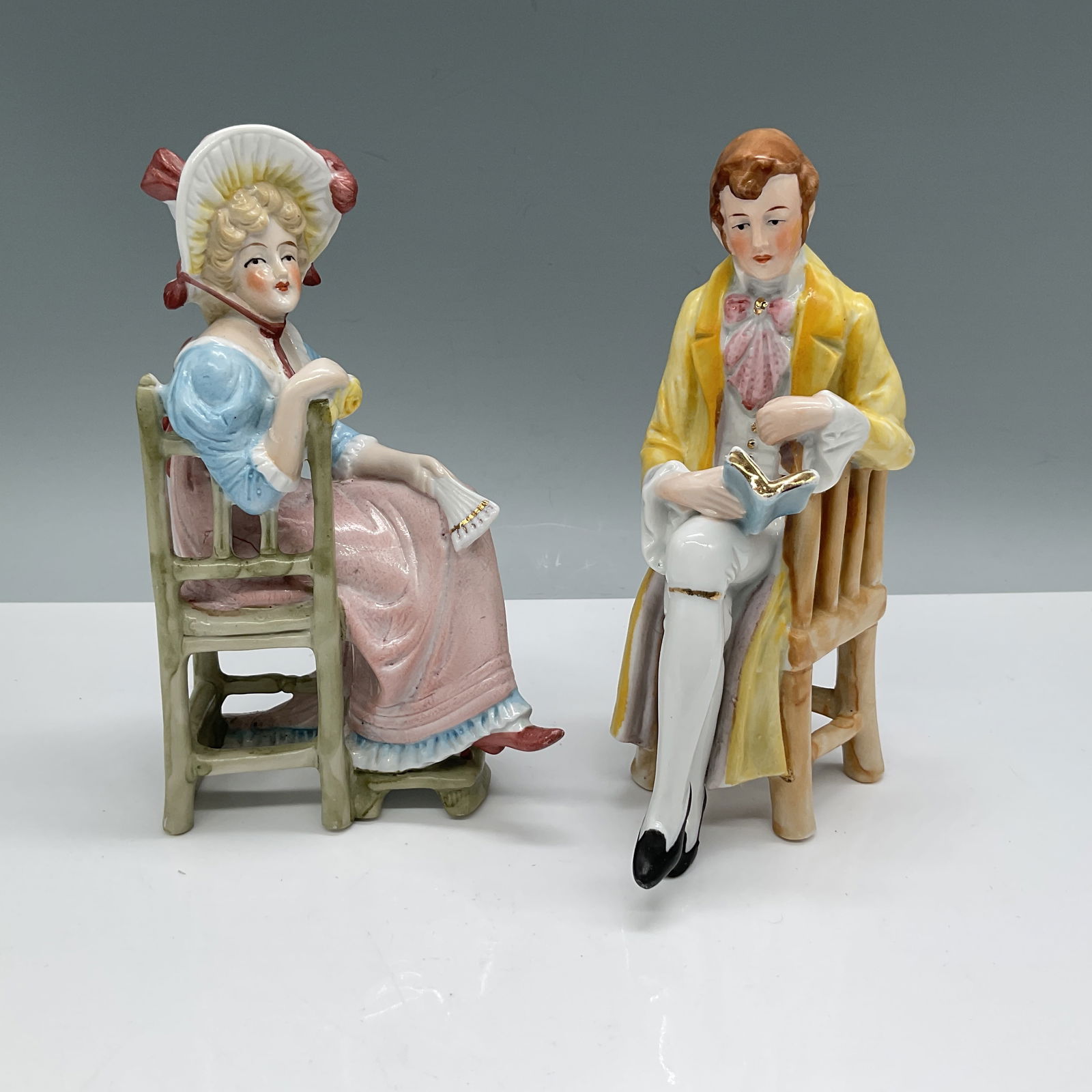 2pc Porcelain Figurine Set, Regency Style Couple: A charming two-piece porcelain figurine set depicting a Regency-era couple seated in wooden-style chairs. The woman wears a pastel pink and blue gown with a bonnet, holding a fan, while the gentleman