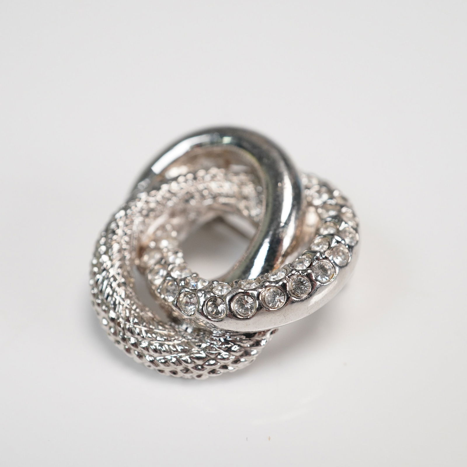 Silver Tone Interlocking Loop Rhinestone Brooch: Silver tone brooch featuring three interwoven loops, each with a unique finish"smooth, braided, and pave set with clear rhinestones. The sculptural design offers a bold yet versatile statement,