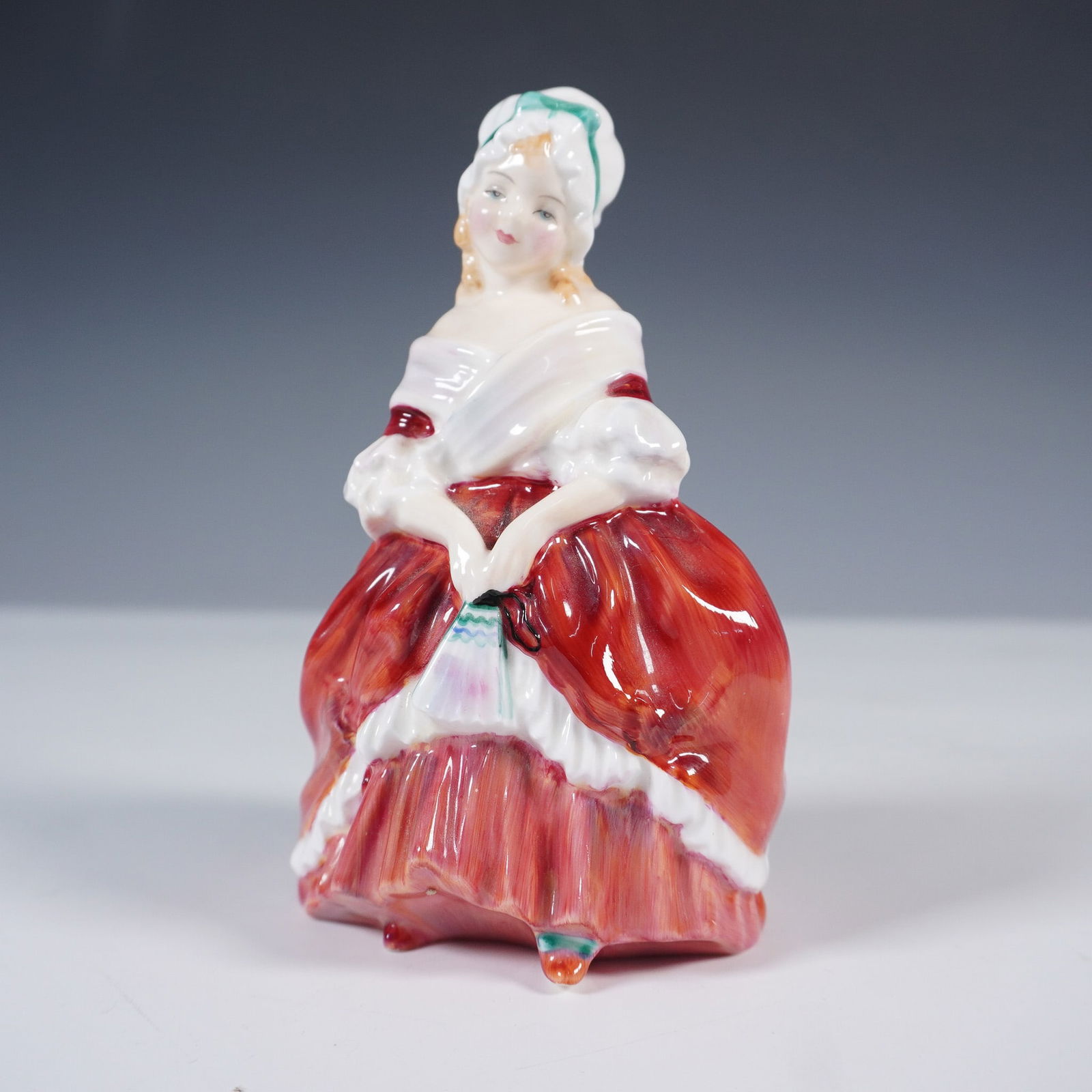 Royal Doulton Figurine, Peggy HN2038: This hand-painted figure depicts a young woman in an 18th-century style dress with vibrant red and white flowing layers and a white cap adorned with a light blue ribbon. The figure features finely mod