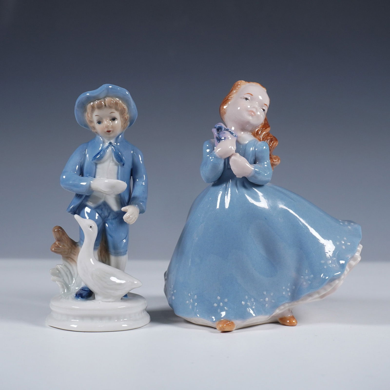 2pc Porcelain Figurines, Holland Blue Boy and Girl: Charming pair of vintage porcelain figurines featuring a girl in a sweeping blue dress marked Holland Molo, and a boy in matching blue attire beside a goose, marked Made in Taiwan. The Holland Molo fi