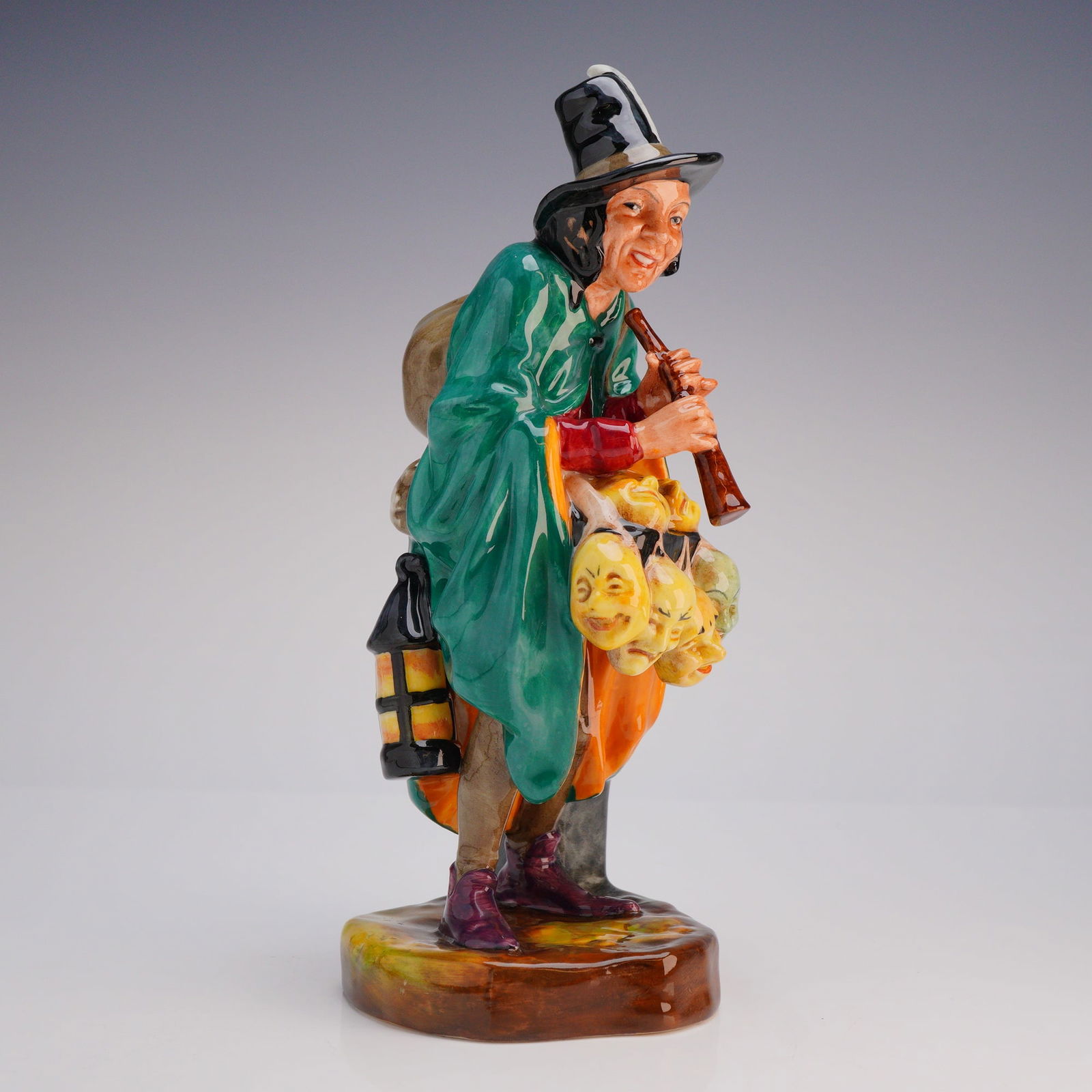 Royal Doulton Porcelain Figurine The Mask Seller HN2103: The figure is attributed to designer Leslie Harradine. The polychrome enameled figure depicts a male street vendor or entertainer, modeled standing while playing a woodwind instrument. He is dressed i