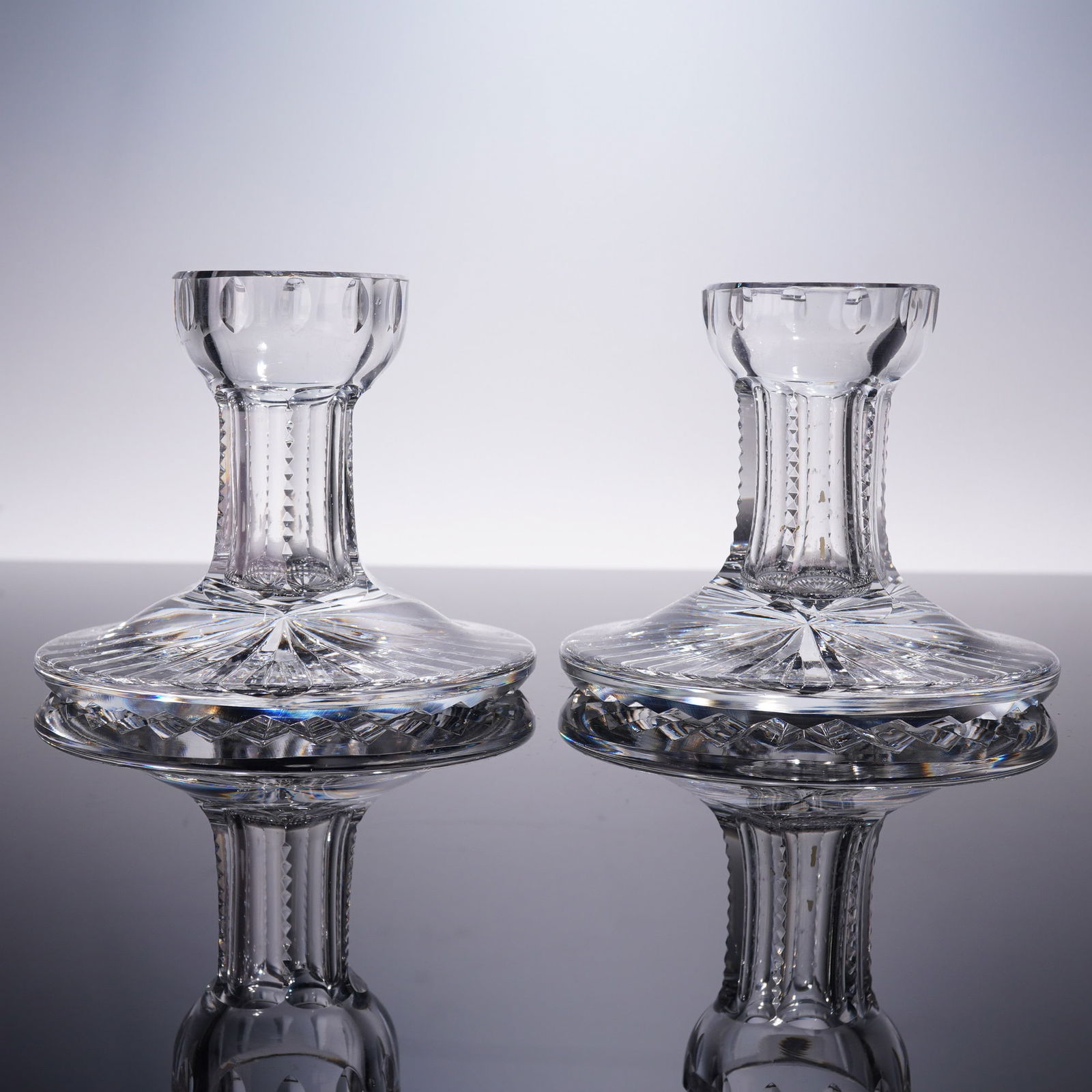 2pc Cut Crystal Candlesticks with Starburst Bases: Pair of cut crystal candlesticks featuring flared, scalloped rims and faceted stems with vertical diamond cuts, leading to broad circular bases with striking starburst patterns. The clear crystal catc