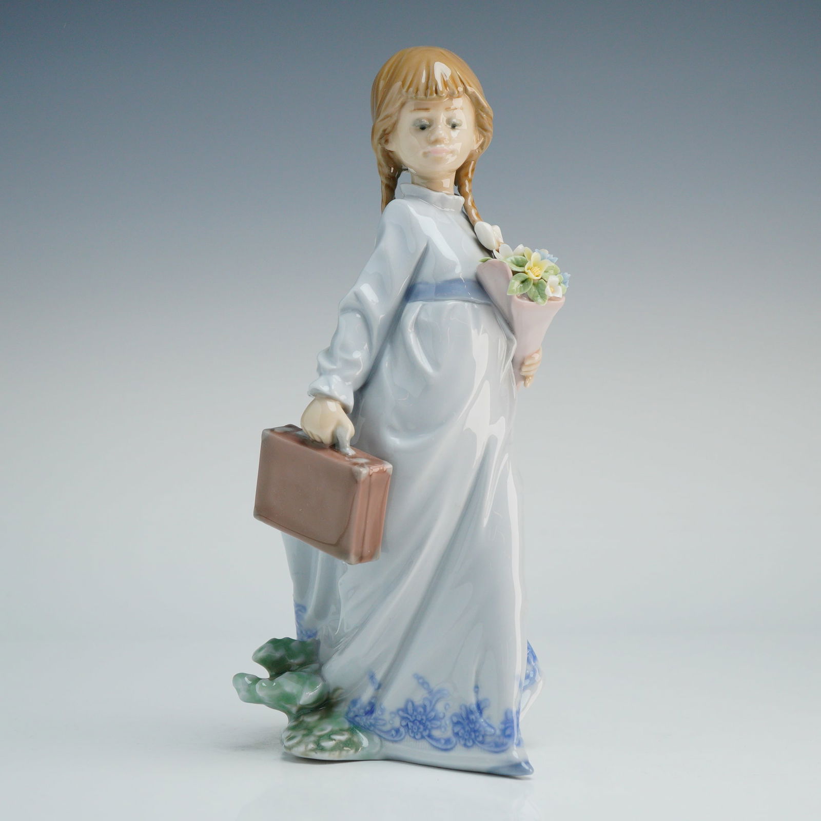 Lladro Porcelain Figurine School Days 1007604: The figure depicts a young girl with blonde braids, carrying a brown suitcase and a bouquet of flowers. The subject is dressed in a full-length pale blue gown with a dark blue floral motif at the hem