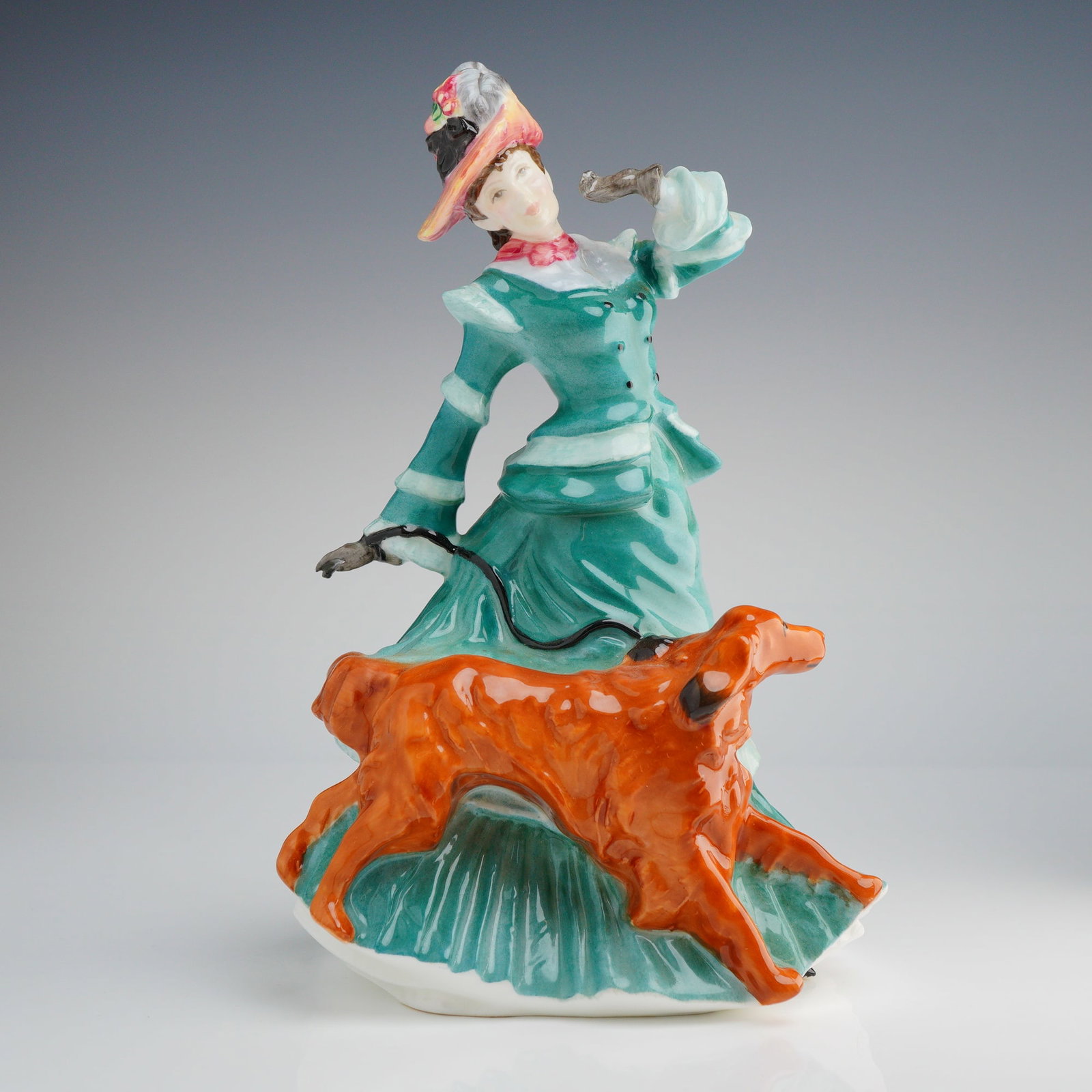 Royal Doulton Figurine, Autumntime HN3621: The piece depicts a woman in a long, high-collared Victorian-style dress, primarily rendered in teal and white, wearing a plumed hat. She is shown walking a reddish-orange sporting dog, identified in