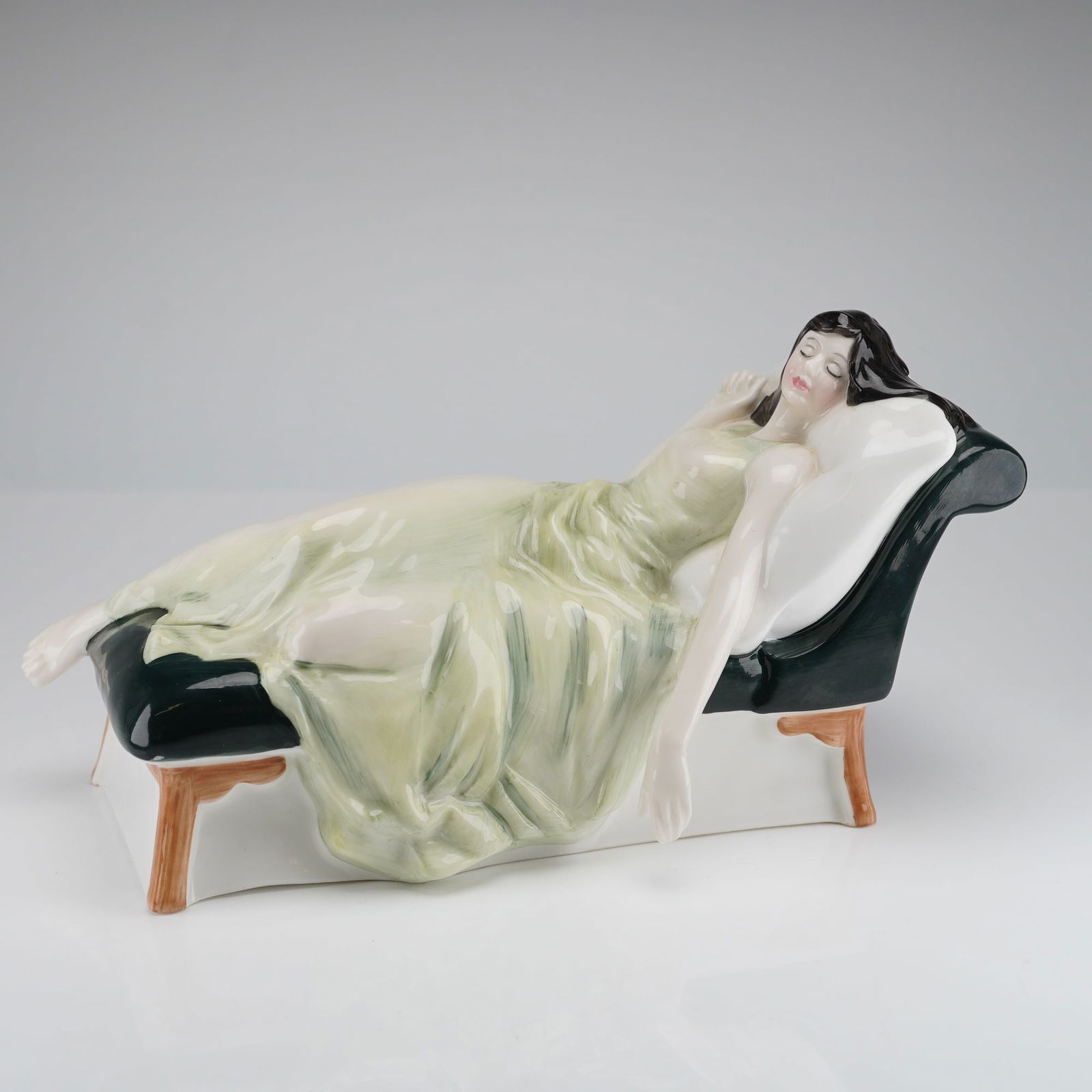 Royal Doulton Figurine Sleeping Beauty HN3079: The figurine depicts a woman with dark hair, clad in a flowing pale green gown, reclining dramatically on a dark green chaise lounge with white cushions and simulated wooden legs. The piece is execute