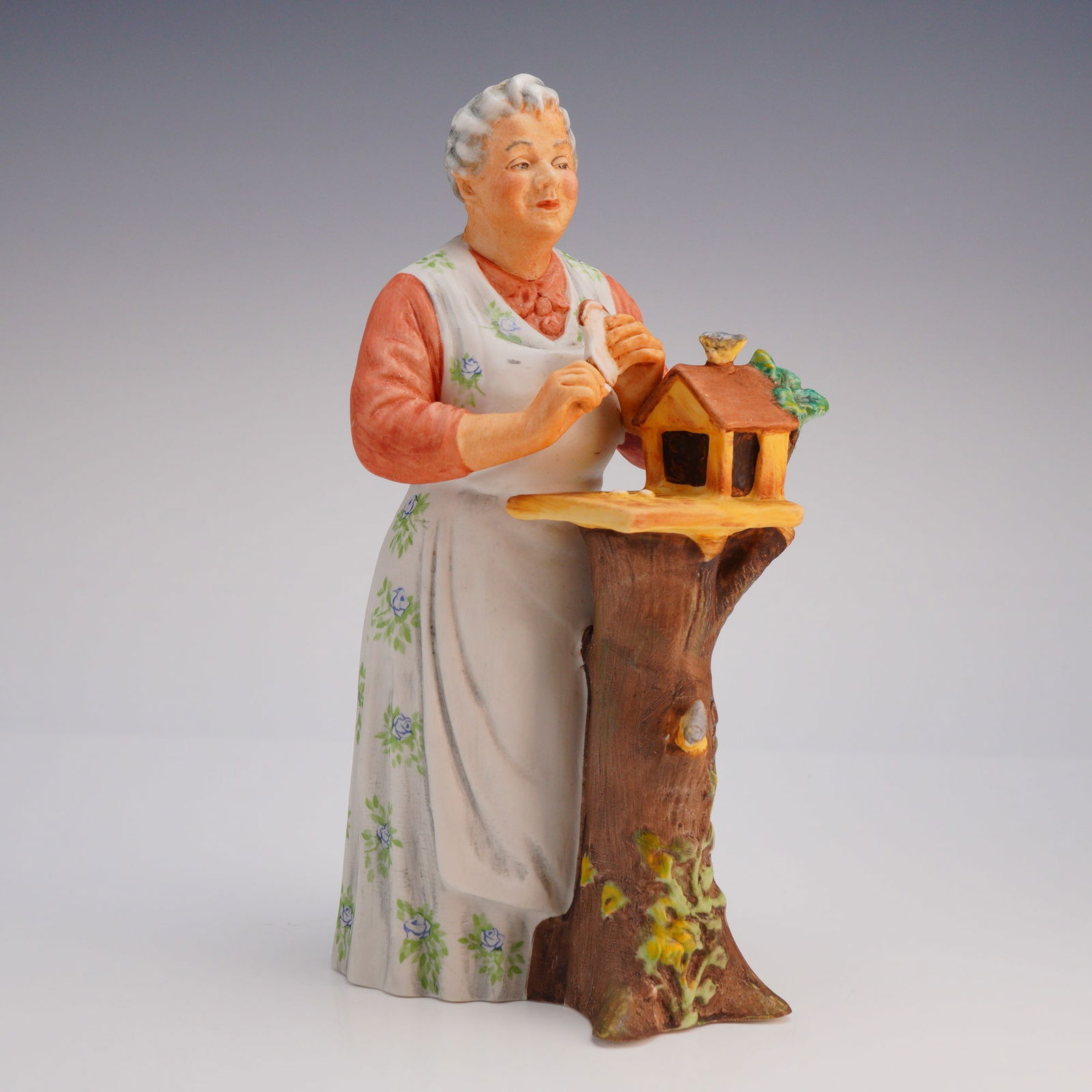 Royal Doulton Porcelain Figurine Good Morning HN 2671: The piece depicts an elderly woman, hand-painted in decorative tones of brown, orange, white, green, and blue, wearing an orange top and a long white apron patterned with small blue flowers and green