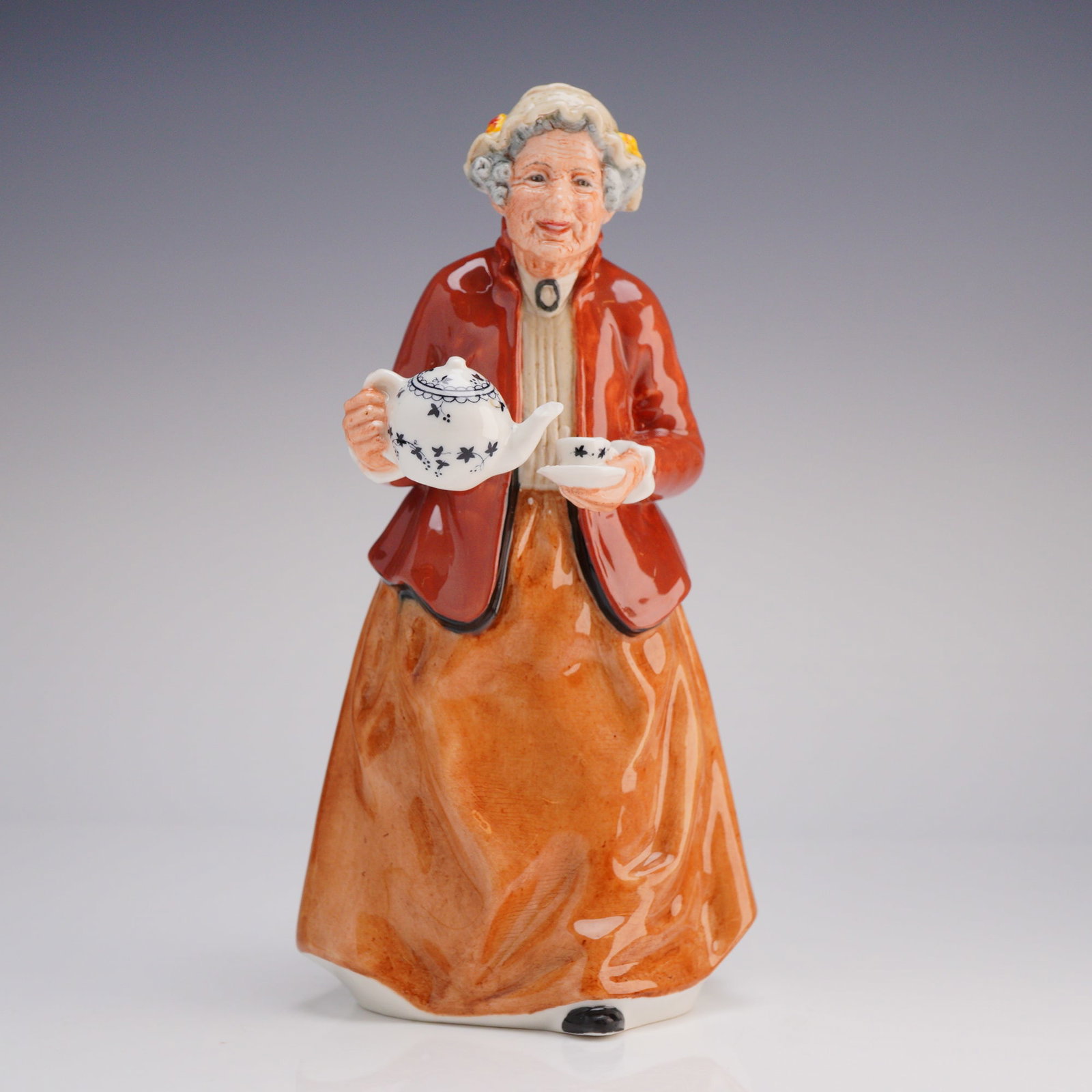 Royal Doulton Figurine Teatime HN 2255: The figurine depicts an elderly woman, often referred to as 'Grandma' in market listings, dressed in a rust-orange skirt and a reddish-brown jacket, preparing to serve tea. She holds a small teapot an