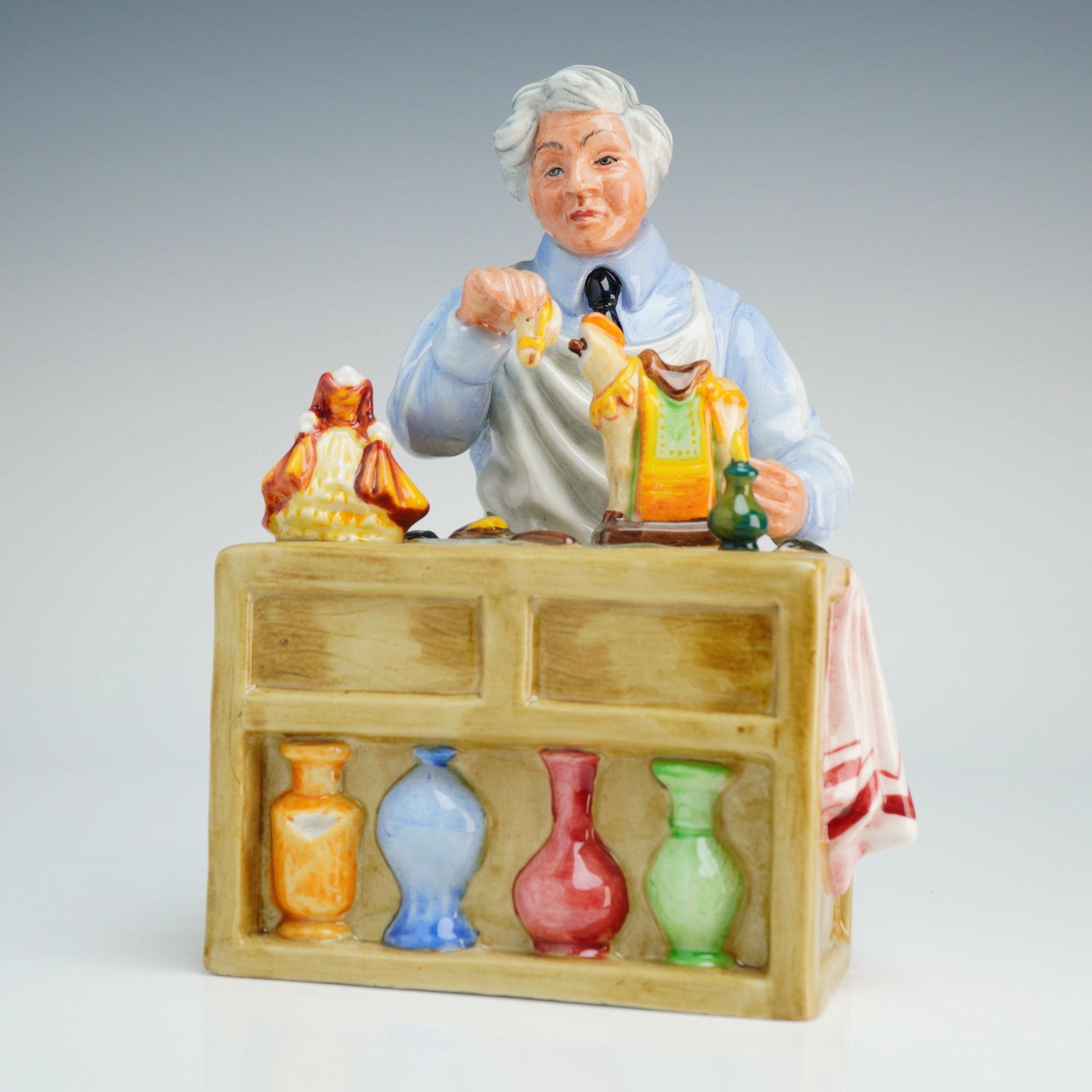 Royal Doulton Figurine The China Repairer HN2943: This hand-made and hand-decorated object is constructed of polychrome porcelain. The scene features an elderly craftsman with white hair seated at a tan workbench, wearing a blue shirt and white apron
