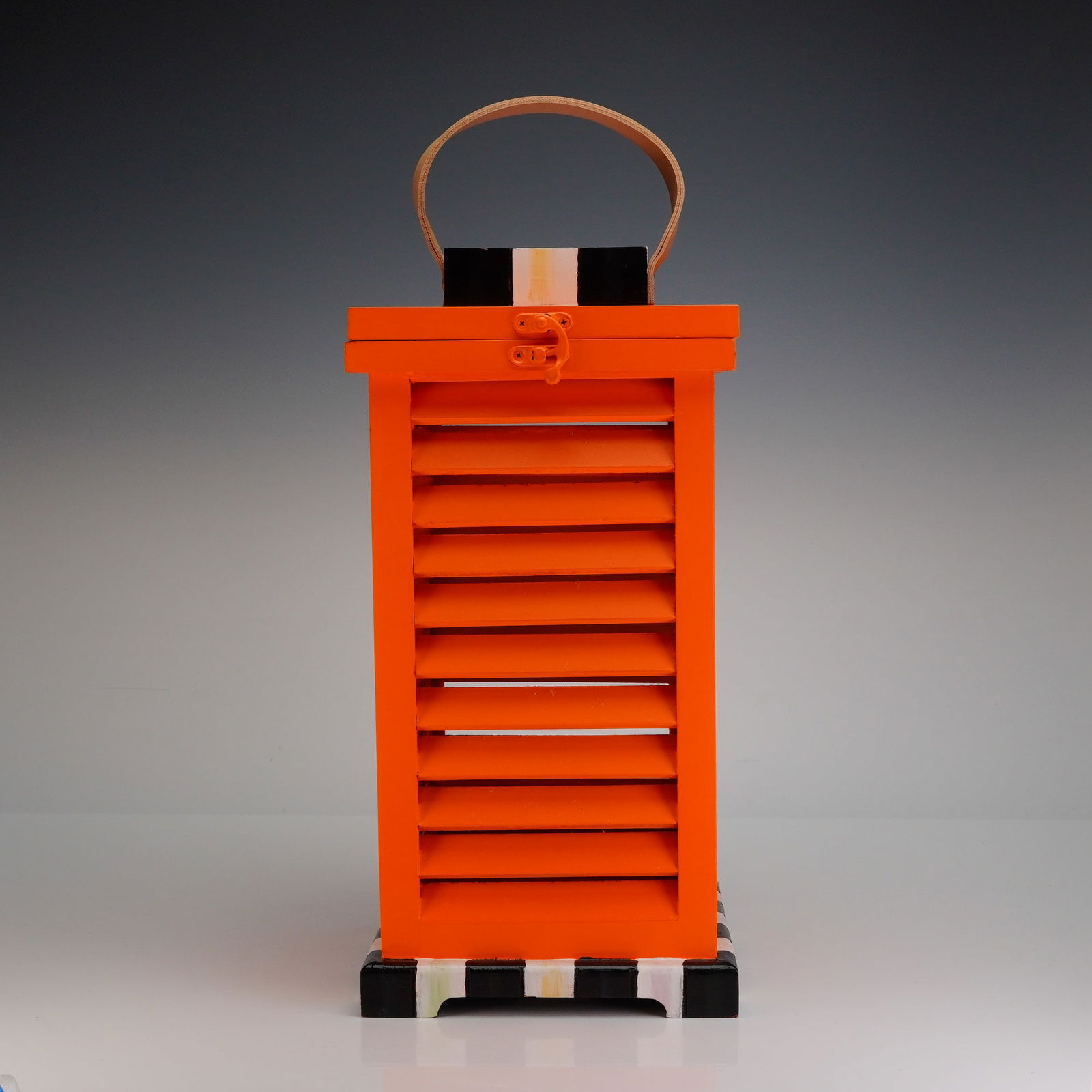MacKenzie Childs Small Orange Shutter Lantern: This decorative piece is constructed primarily of painted wood in a contemporary style. The object features a tall, rectangular body with slatted sides finished in a vibrant, high-gloss orange paint.