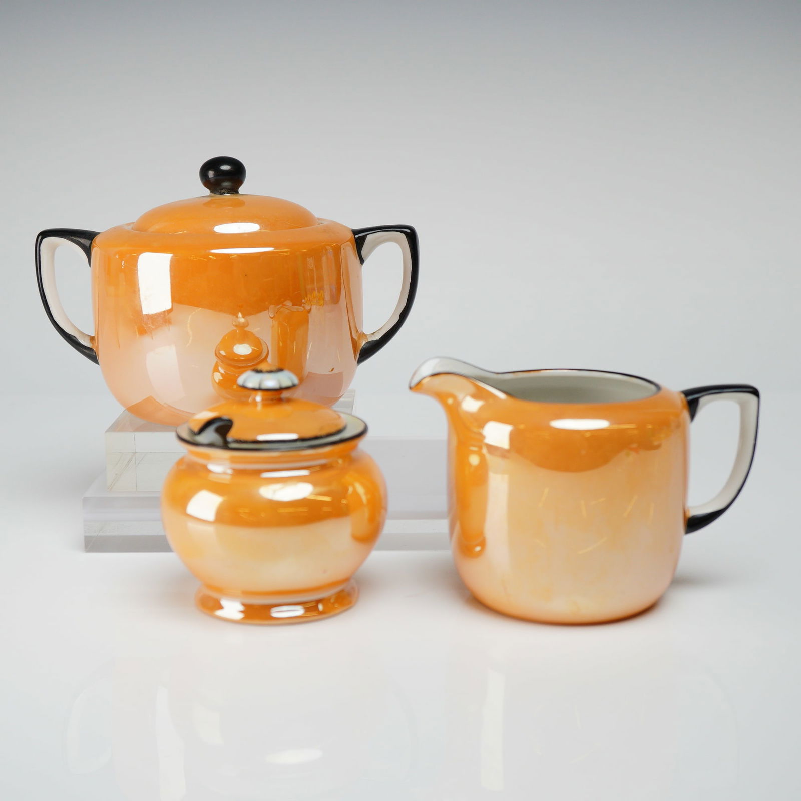 3pc Noritake Orange Lusterware Creamer, Sugar with Lid & Jam Jar Set: The porcelain pieces feature a vibrant iridescent orange or peach luster glaze, characteristic of early to mid-20th-century Japanese export ware. The handles, rims, and finials are sharply contrasted