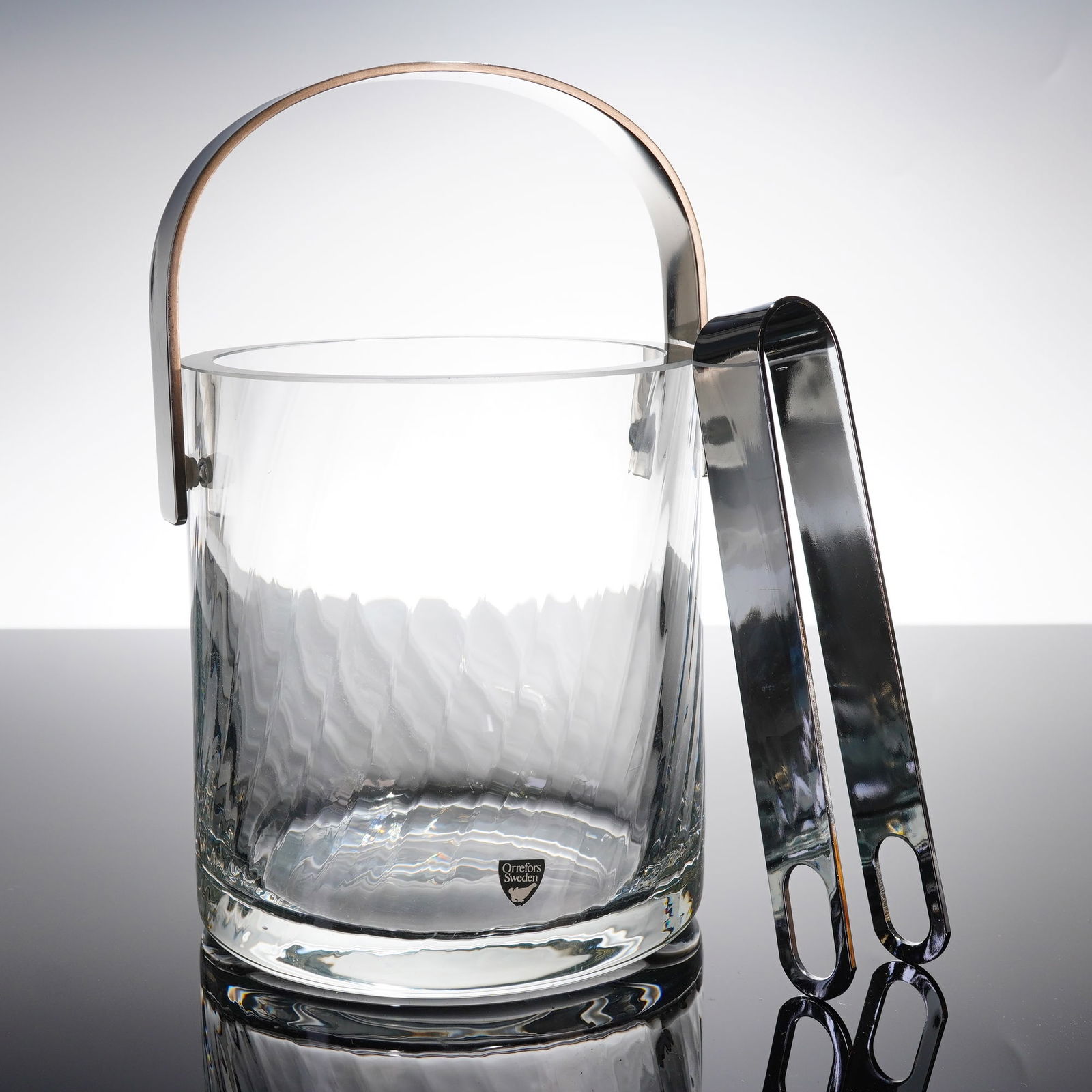 Orrefors Crystal Ice Bucket and Tongs Set: The set consists of a thick-walled cylindrical ice bucket made of clear glass, a removable stainless steel bail handle, and a pair of matching stainless steel tongs. The glass bucket features a swirli