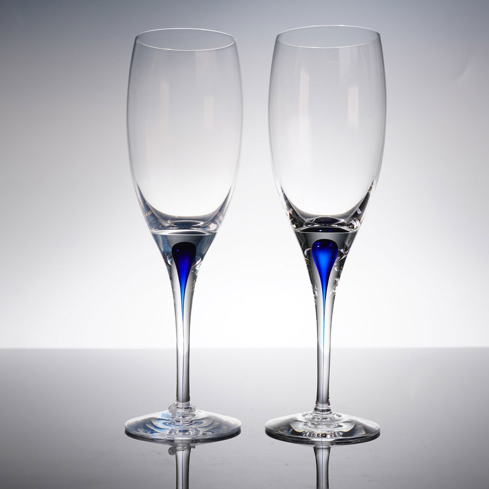 A Pair of Orrefors Intermezzo Blue Champagne Flutes: A pair of contemporary champagne flutes manufactured by Orrefors in the Intermezzo Blue patern. Each flute is crafted from clear glass featuring a tall, tapered bowl supported by a slender stem and ci