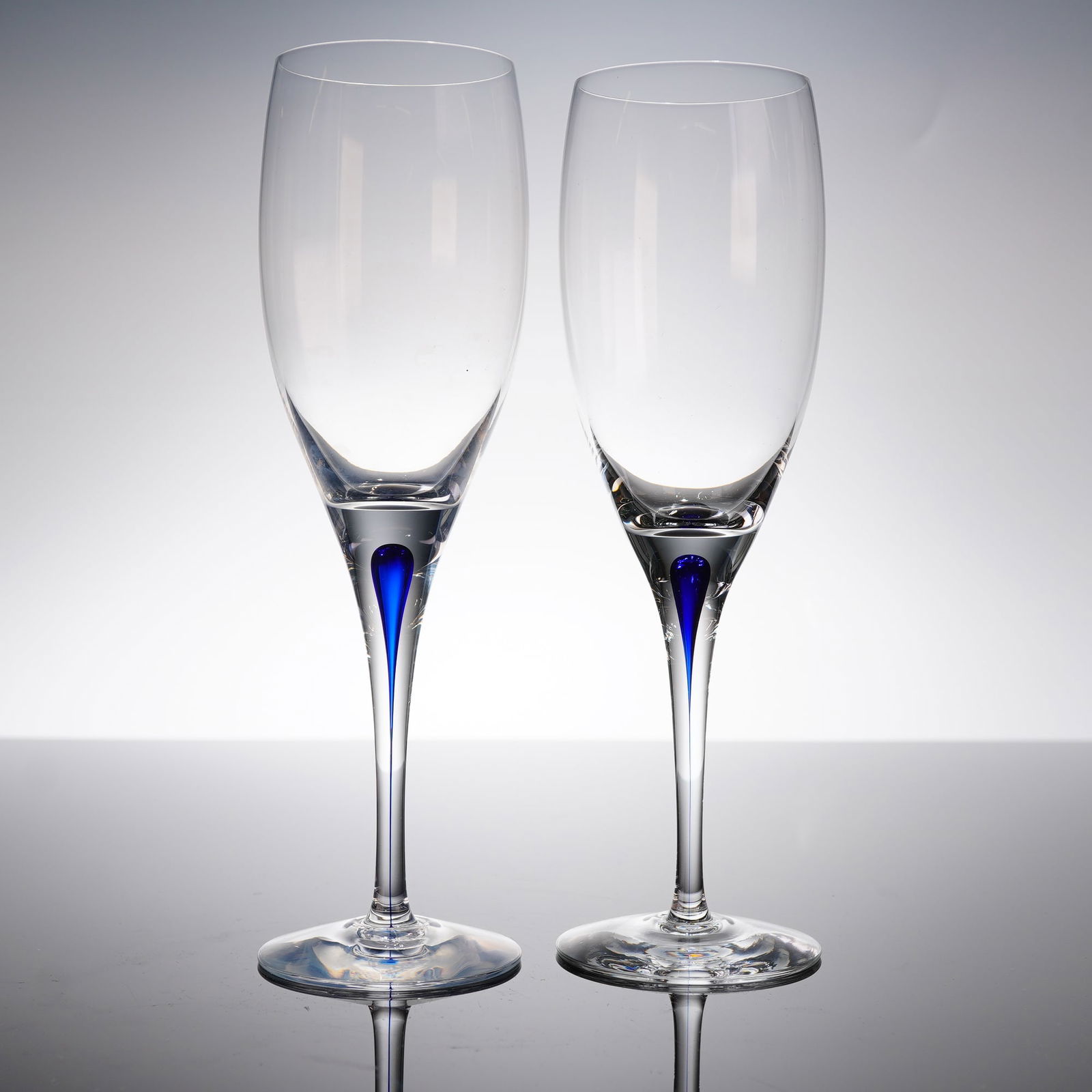 A Pair of Orrefors Intermezzo Blue White Wine Glasses: A pair of contemporary clear champagne flutes in the Intermezzo Blue patern. Each glass features a tall, profile, a narrow stem, and a round foot. The distinguishing characteristic is the internal inc