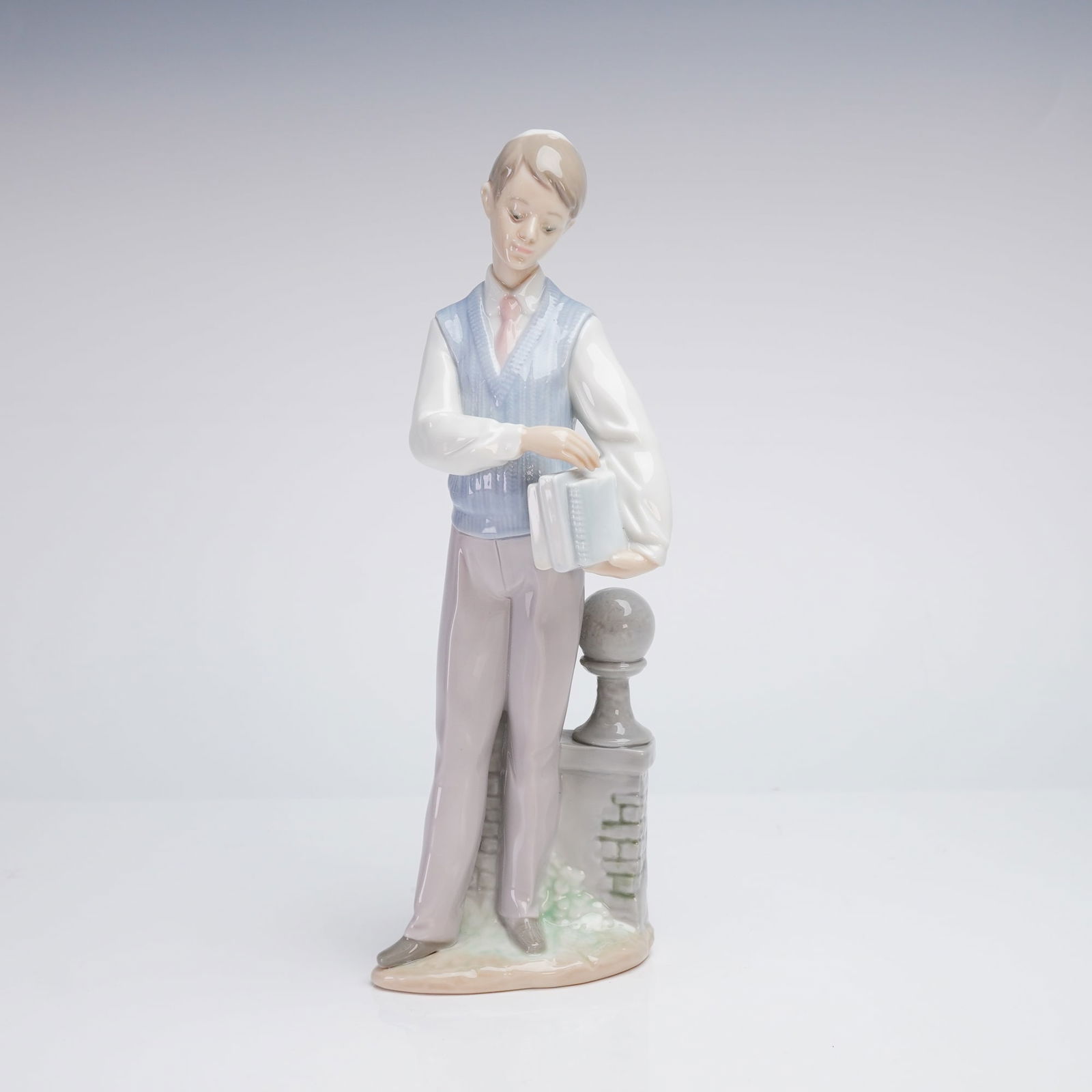 Lladro Hebrew Scholar Porcelain Figurine 6029: A Lladro porcelain figurine titled Hebrew Scholar, model number 6029. This glazed porcelain sculpture depicts a young male student wearing a yarmulke, blue sweater vest, and pink tie, holding books wh