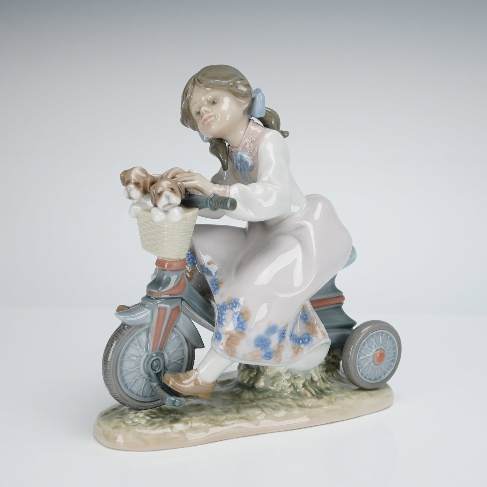 Lladro Traveling in Style Figurine 5680: A single Lladro Porcelain Figurine titled "Traveling In Style," model number 01005680. The glazed porcelain sculpture depicts a young girl with brown hair and a blue ribbon riding a gray and red tricy