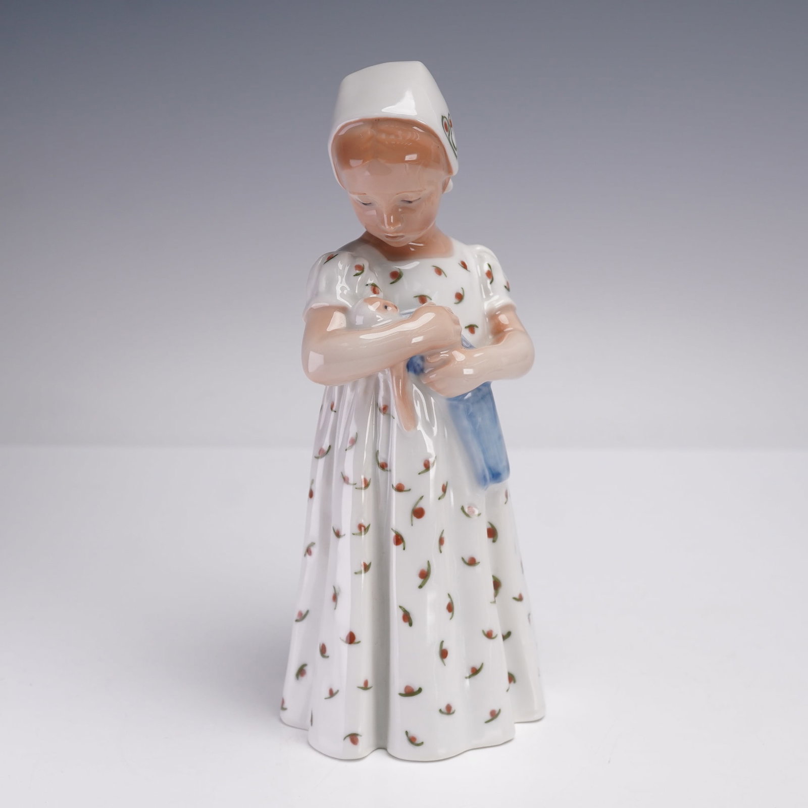 Royal Copenhagen Figurine, Mary with Doll 561: Charming porcelain figurine depicting a young girl in a long white dress patterned with delicate red and green floral sprigs. She wears a matching white kerchief tied with a bow at the back, and she t